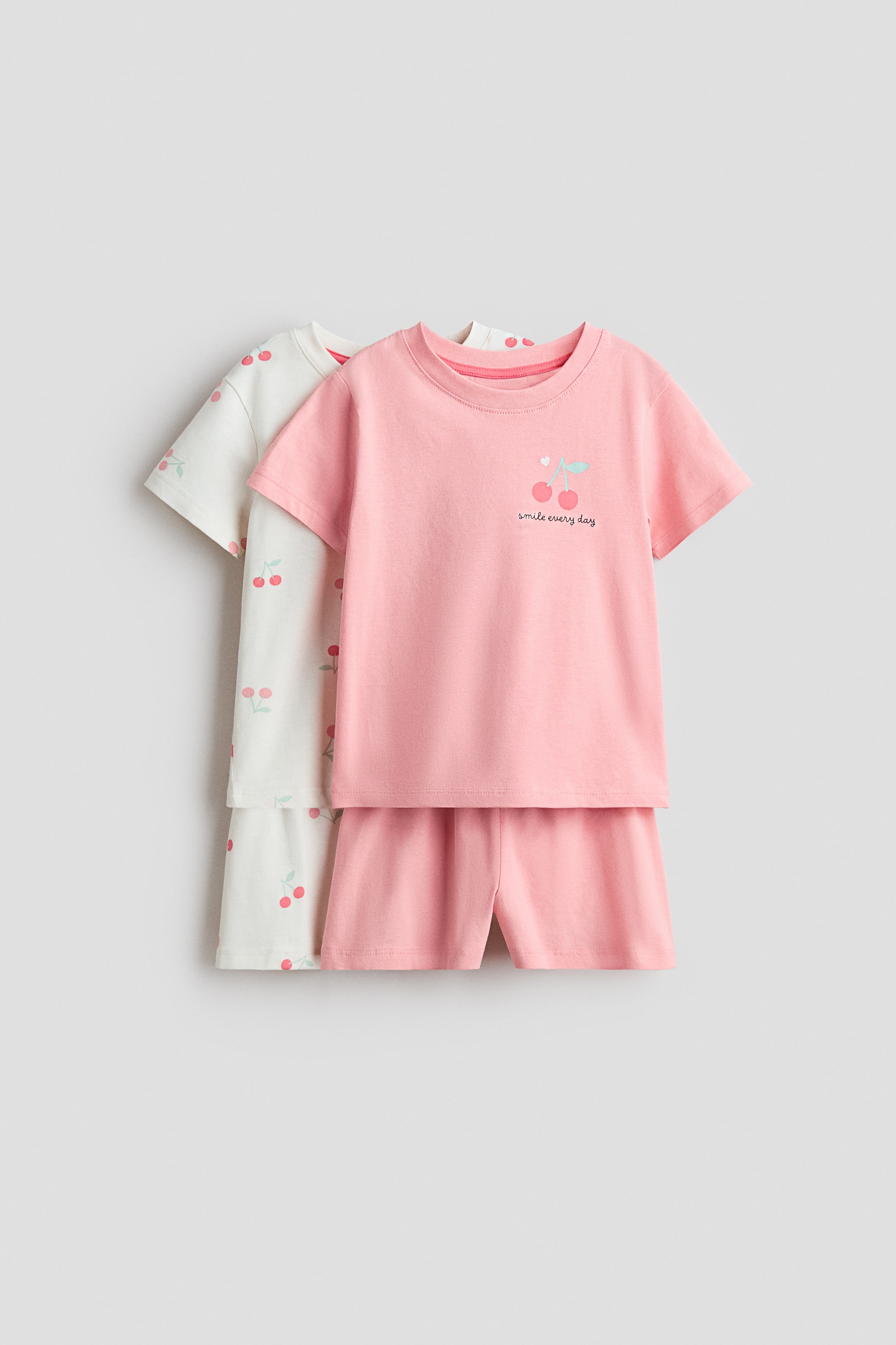 View larger image: The pyjamas showcase two sets, each comprising a t-shirt and matching shorts, both facing front. The front set highlights a pink t-shirt with a ribbed neckline, gently dropped shoulders, and a cherry graphic with the text "smile every day," paired with matching pink shorts featuring covered elastication at the waist. Partially visible behind it is a second set, featuring a white t-shirt and shorts, both adorned with an all-over cherry print.