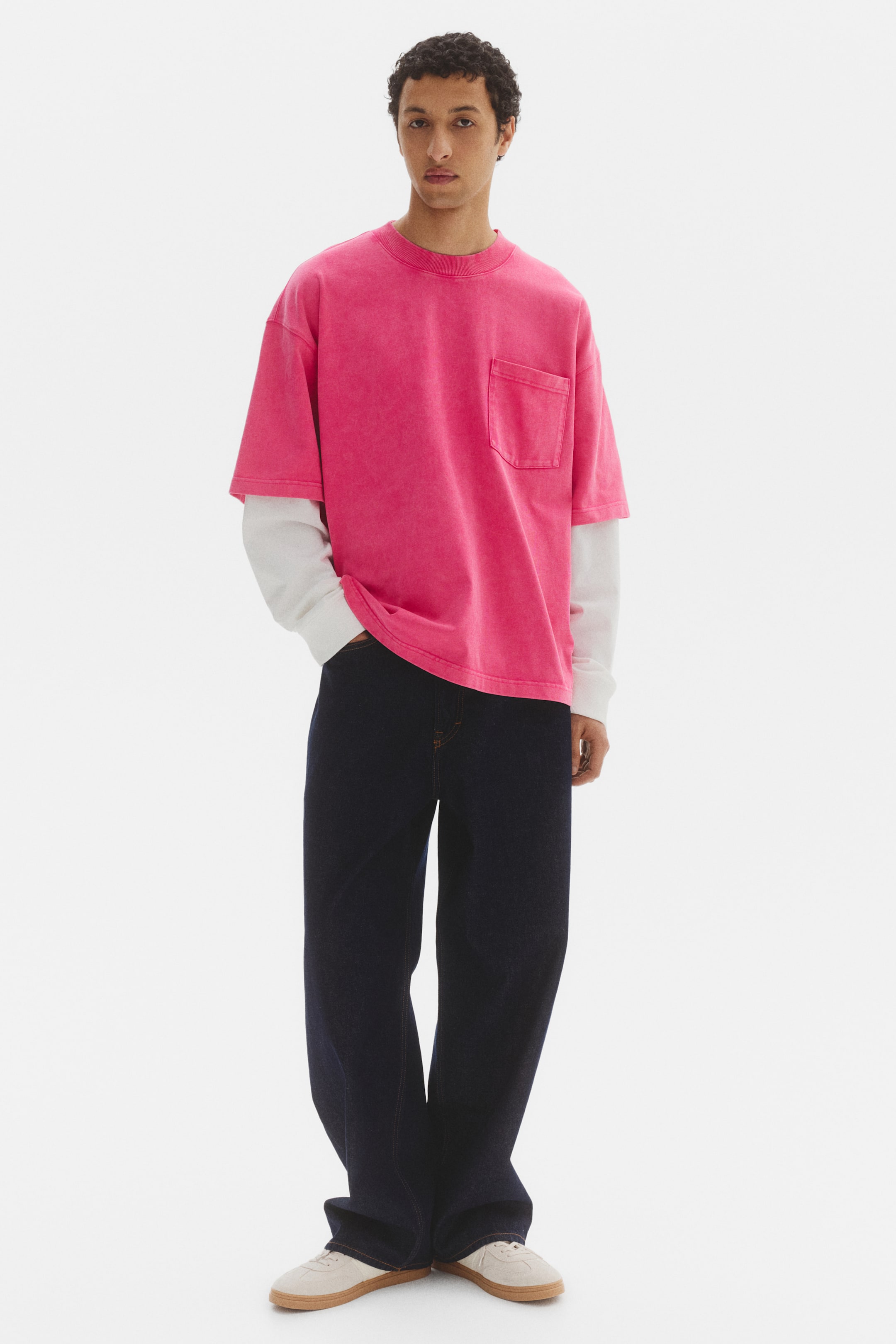 View larger image: "A fuchsia pink oversized t-shirt is facing forward, featuring a crew neck and a patch pocket on the left chest. It is being styled over a white long-sleeve shirt, and its fabric is displaying a faded finish."