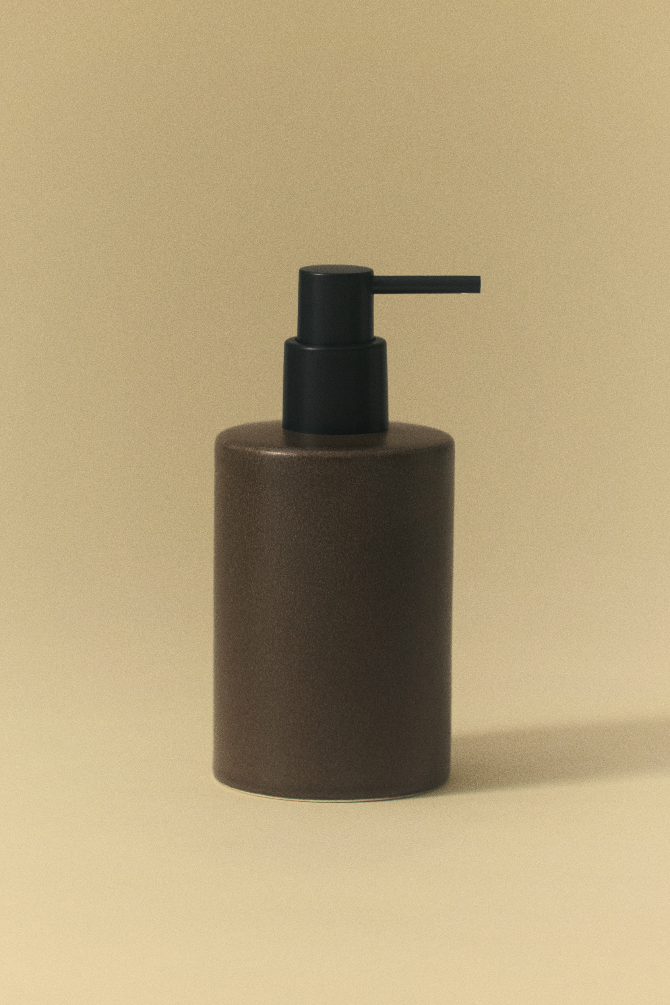 Un distributore di sapone cilindrico è posizionato frontalmente, caratterizzato da un corpo in gres marrone maculato e smaltato con una pompa in plastica nera opaca.