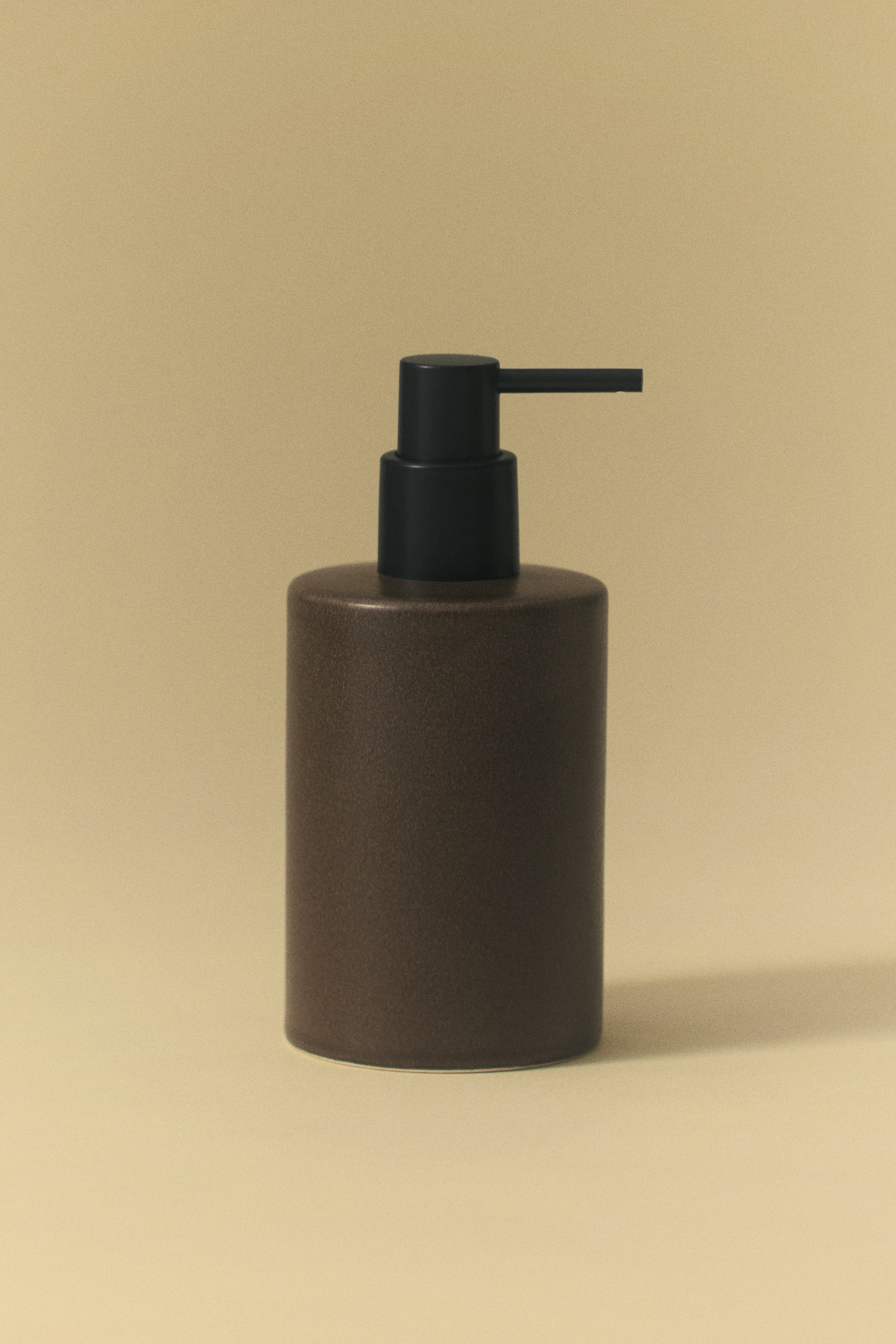 View larger image: A cylindrical soap dispenser is shown from a front-facing angle, featuring a dark brown glazed stoneware body with a subtle speckled finish. It is topped with a matte black plastic pump that has a straight, horizontal nozzle.