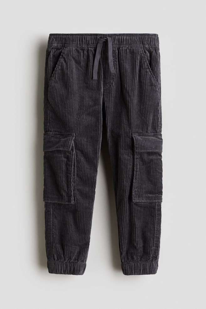 Vorderansicht von dunkelgrauen Cordhosen, die einen elastischen Bund mit sichtbarem Kordelzug aufweisen. Die Hosen zeigen zwei seitliche Schlitztaschen, zwei große Klapp-Cargotaschen an den äußeren Oberschenkeln und gerippte Knöchelbündchen. - 1