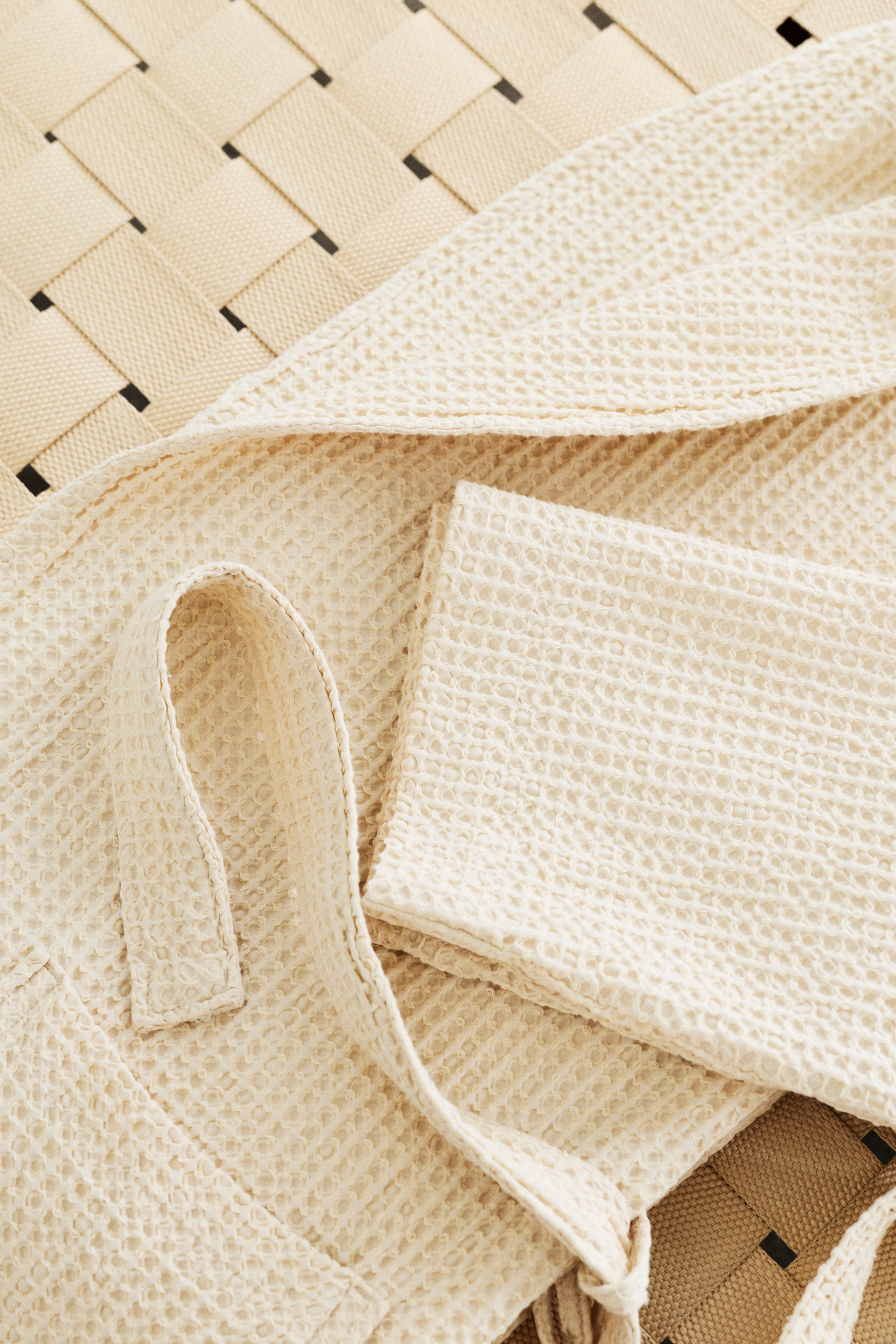 Zobraziť väčší obrázok: Krémový úpletový odev je rozložený, ukazujúci svoju textúrovanú povrchovú úpravu. Viditeľný je pásik na zaväzovanie spolu s malou textilnou slučkou.