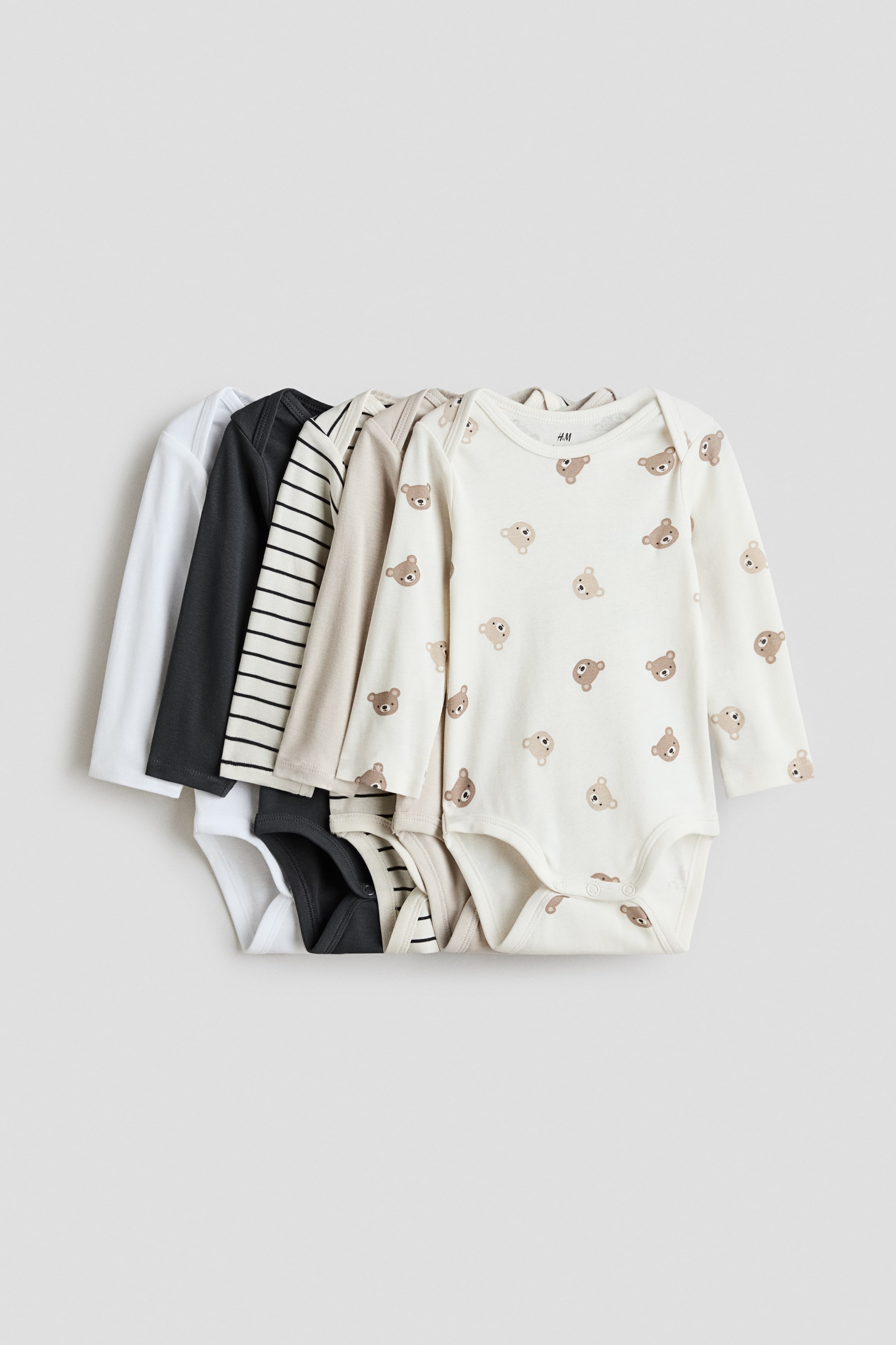 View larger image: Long-sleeved bodysuits are showcasing their FRONT view in a stack of five, featuring lapped shoulders and press-studs at the crotch. The bodysuits are available in solid white, dark grey, cream with black stripes, solid light beige, and off-white with an all-over brown bear face pattern.