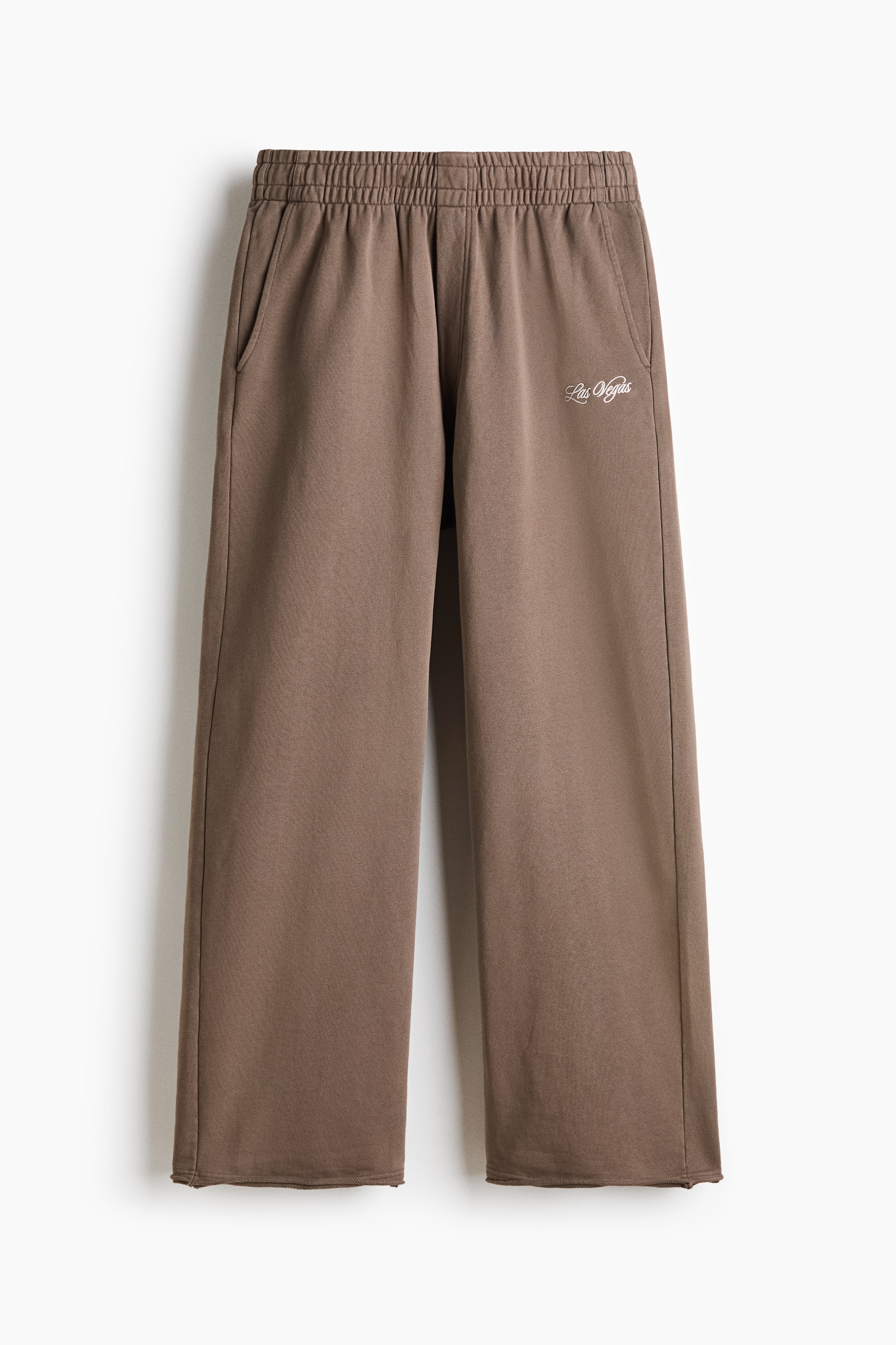 Brown oversized track pants are worn by the model, featuring a relaxed silhouette, a drawstring waistband, and a white script print on the upper left thigh. The model is standing with one hand in a side pocket, styling the pants with a matching brown sweatshirt, an olive jacket, and tan boots.