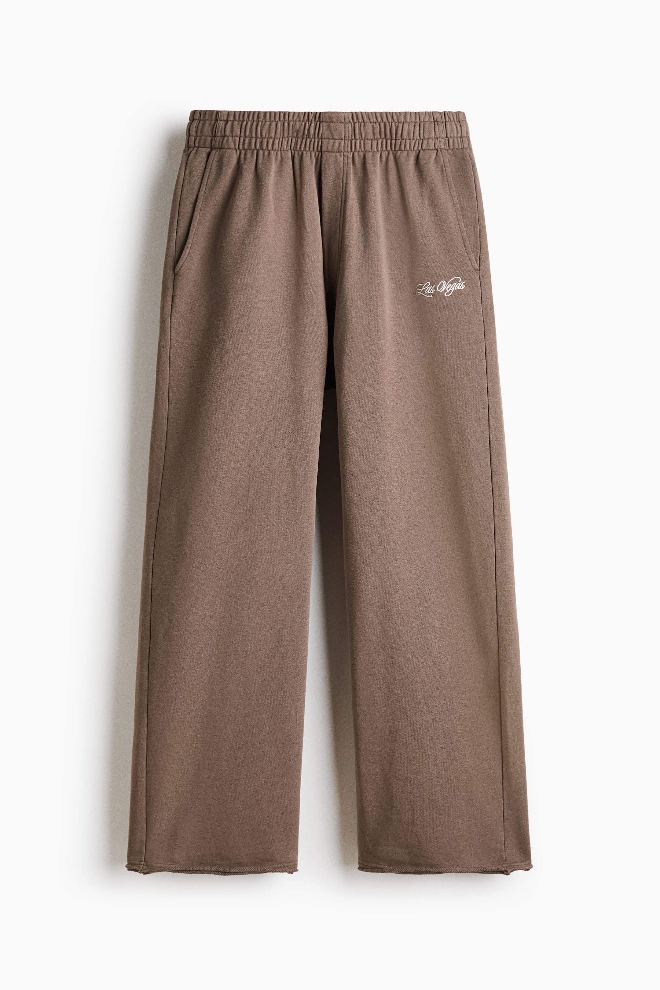 Voir une image plus grande: Une paire de pantalons de sport marron à jambe large est présentée de face, avec une silhouette décontractée et ample, une taille élastique smockée et des poches latérales. Les pantalons affichent un imprimé cursif blanc indiquant « Las Vegas » sur la cuisse gauche et se terminent par des ourlets bruts à bord roulé.