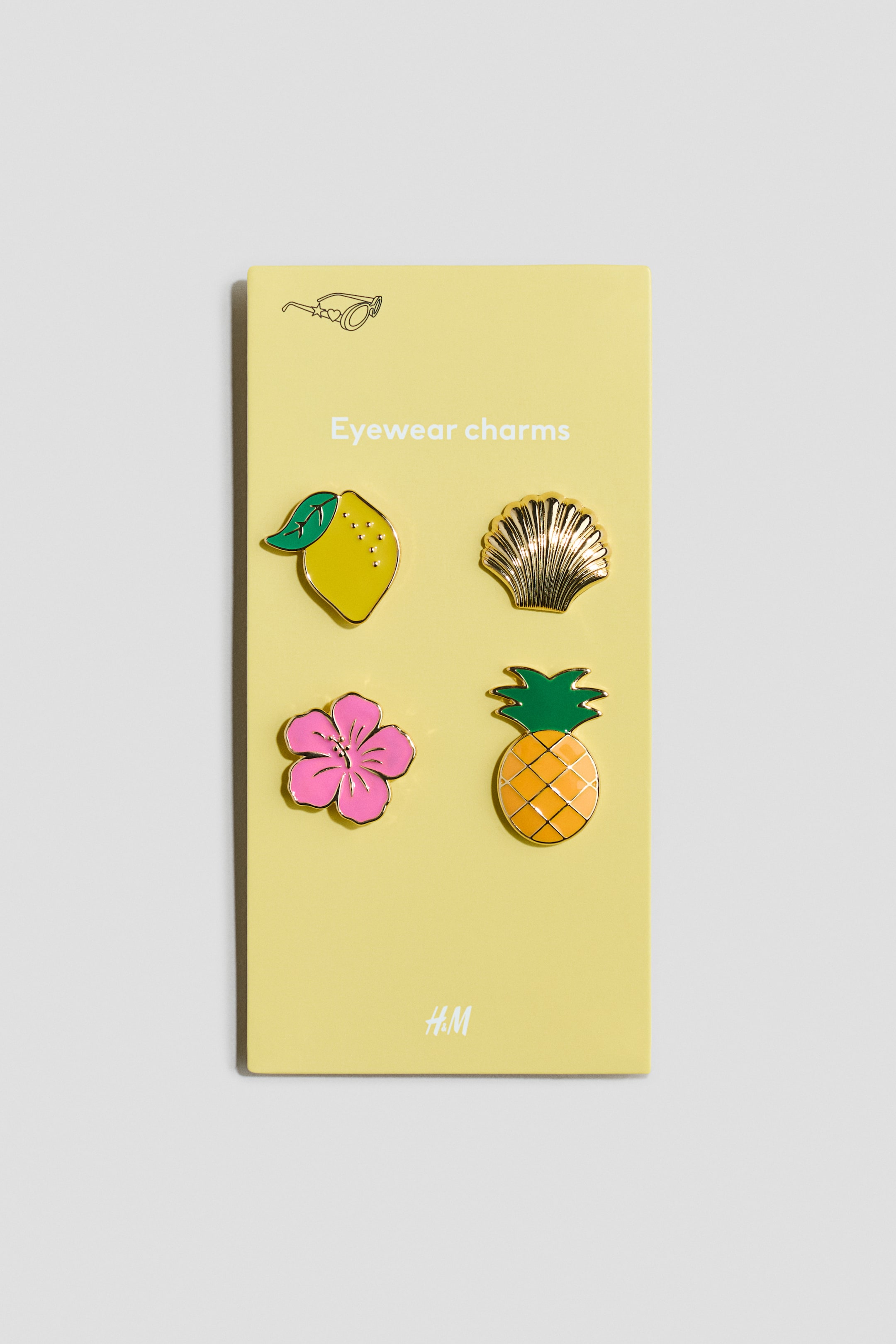 View larger image: Four eyewear charms are displayed on a yellow card, each facing forward. The charms showcase a yellow lemon with a green leaf, a gold scallop shell, a pink flower, and an orange pineapple with green leaves, all bordered in gold. The card also features the text "Eyewear charms" and an eyeglasses graphic at the top, with "H.M" at the bottom.