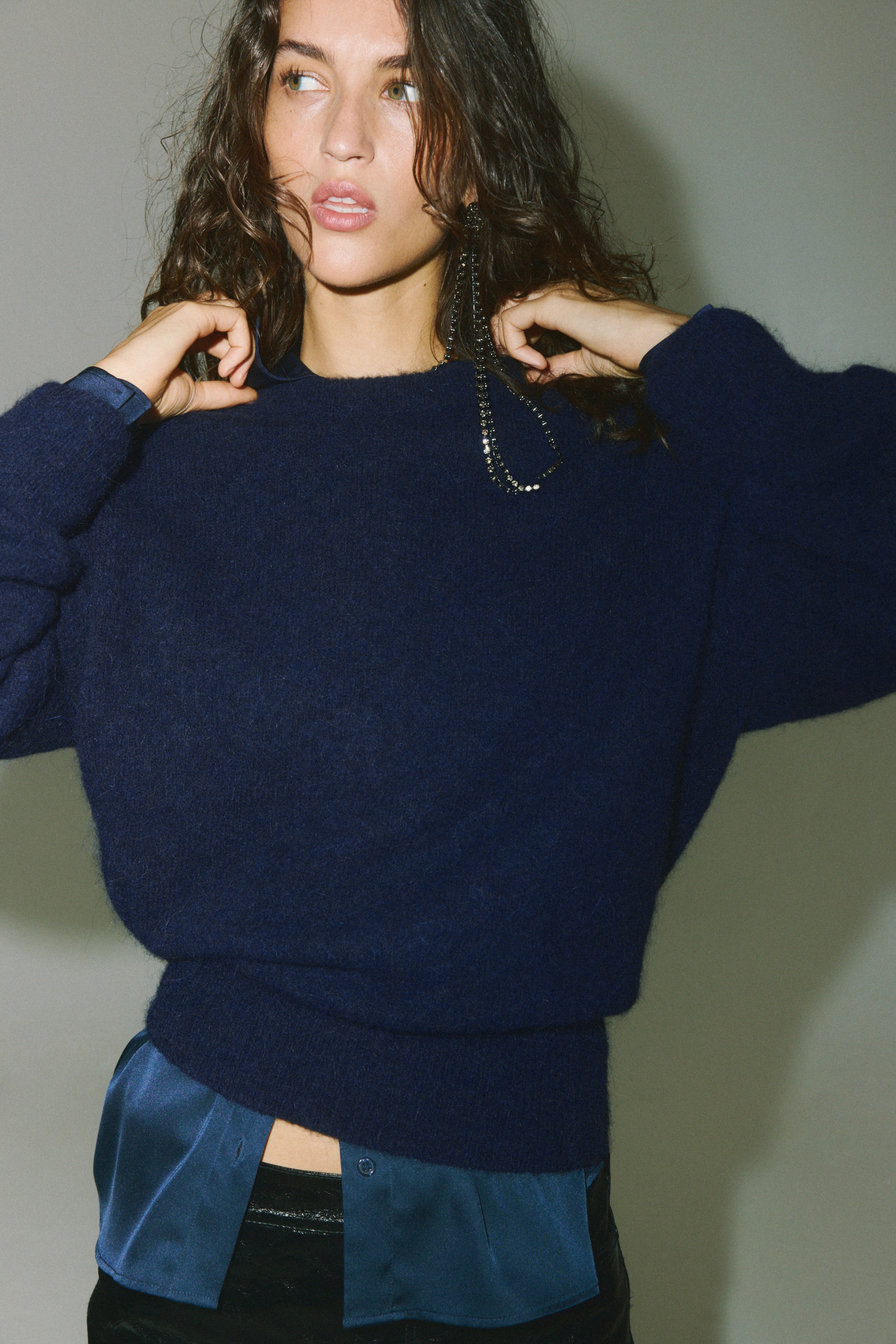 Visualizza immagine più grande: Un maglione oversize in morbida maglia pelosa di colore blu scuro è indossato dalla modella, che sta sistemando il girocollo con entrambe le mani. Il maglione presenta spalle scese e orlo a costine, ed è indossato sopra una camicia in raso blu visibile al colletto e sull’orlo inferiore.