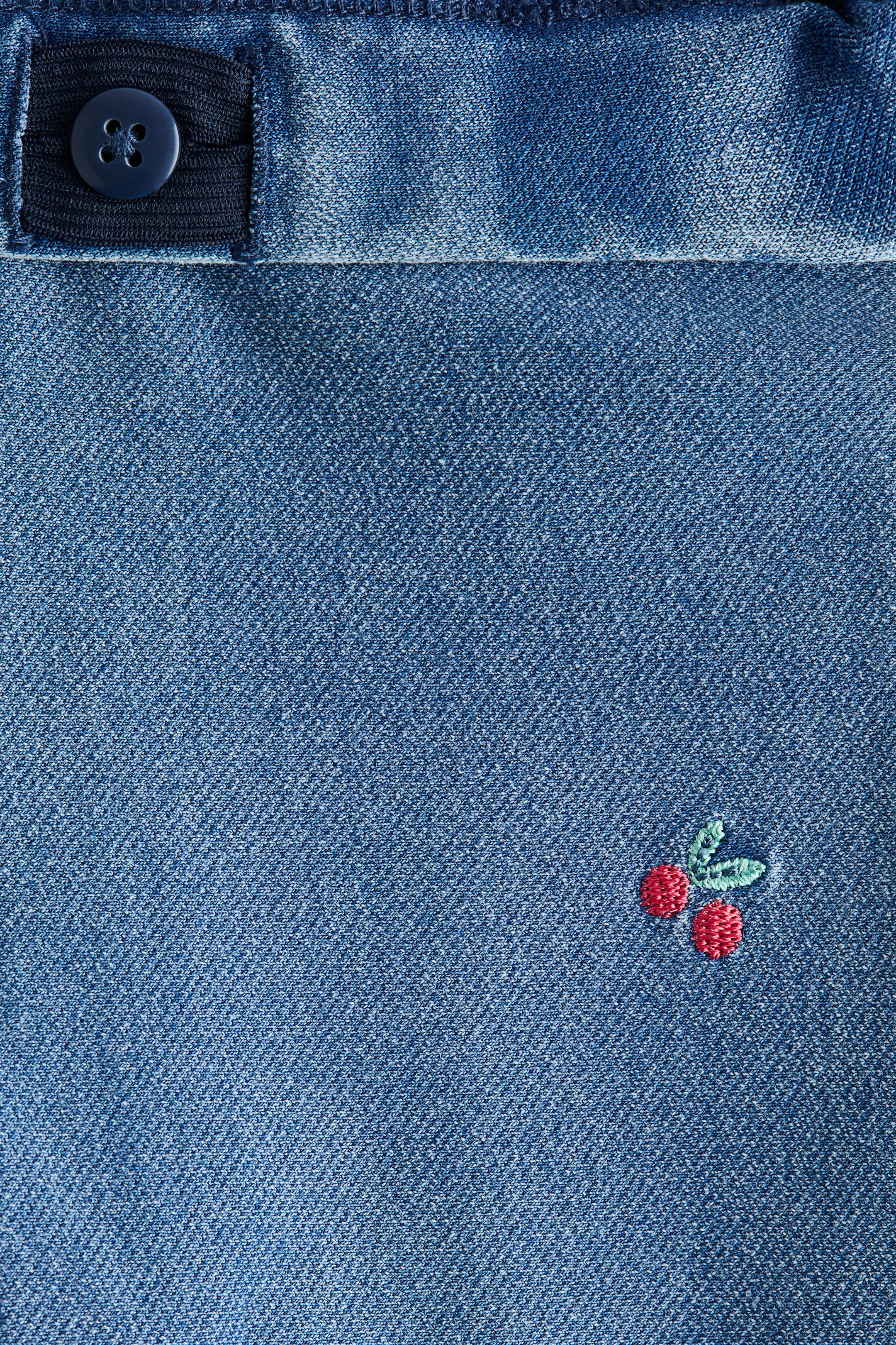 Ampliar la imagen: La tela ligera de la sudadera de los jeggings con apariencia de mezclilla presenta una elasticidad ajustable en la cintura con un botón azul oscuro en la parte superior. Un pequeño diseño bordado de dos cerezas rojas con hojas verdes es visible en la tela.