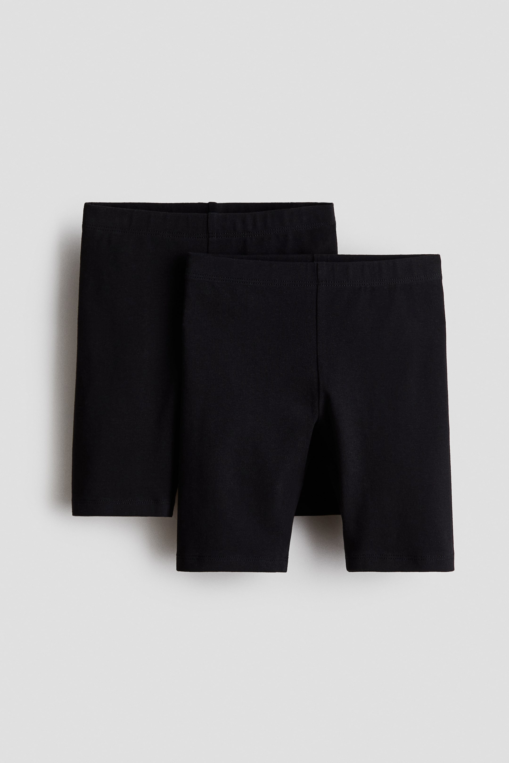 View larger image: Two black cycling shorts lie flat, with one pair partially overlapping the other in a front-facing presentation. The shorts feature smooth, elastic waistbands and extend to mid-thigh length.