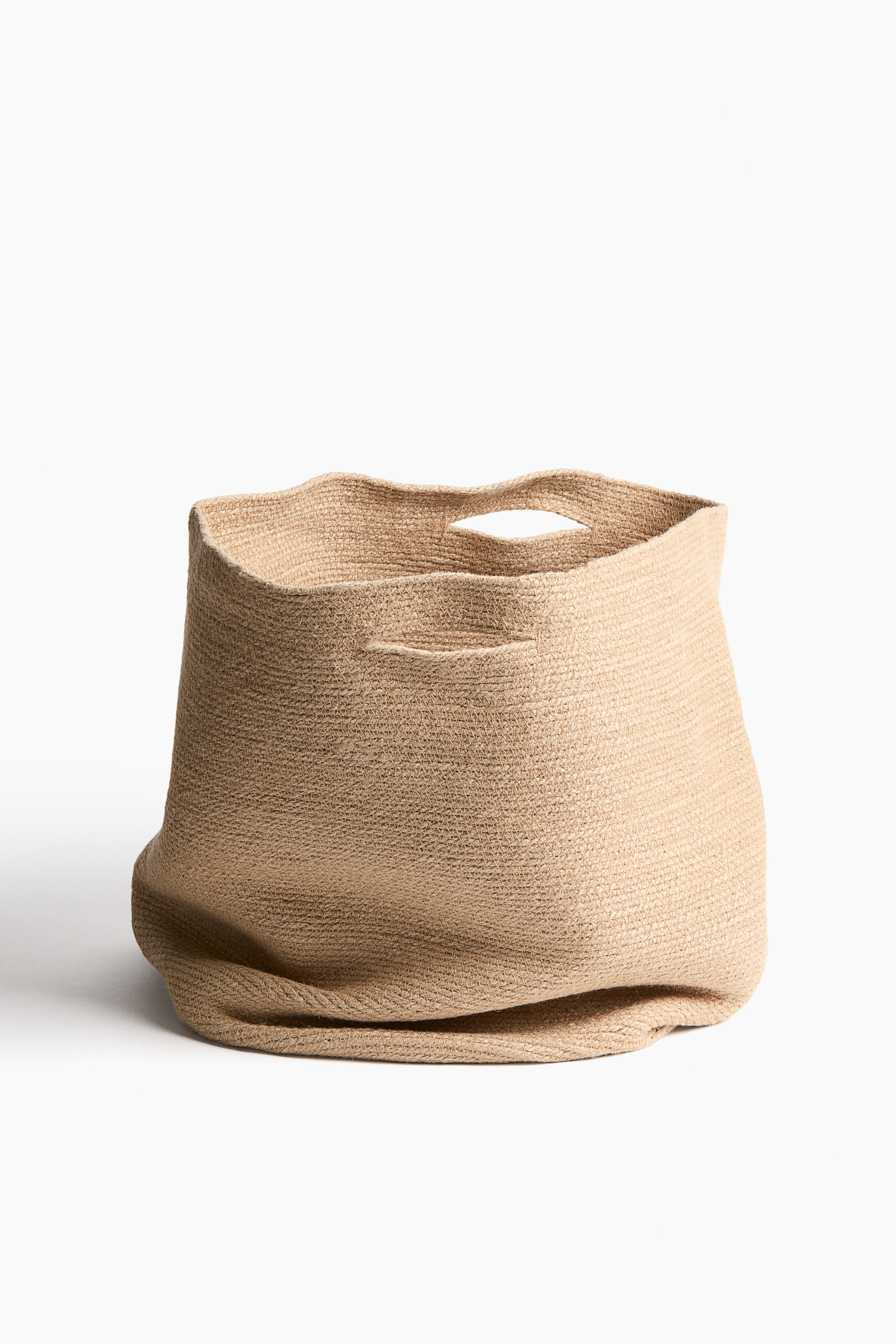 View larger image: A jute storage basket is shown from the front, featuring cut-out handles. Its flexible sides show a relaxed, slouched shape toward the base, while the top edge forms a gentle wave.