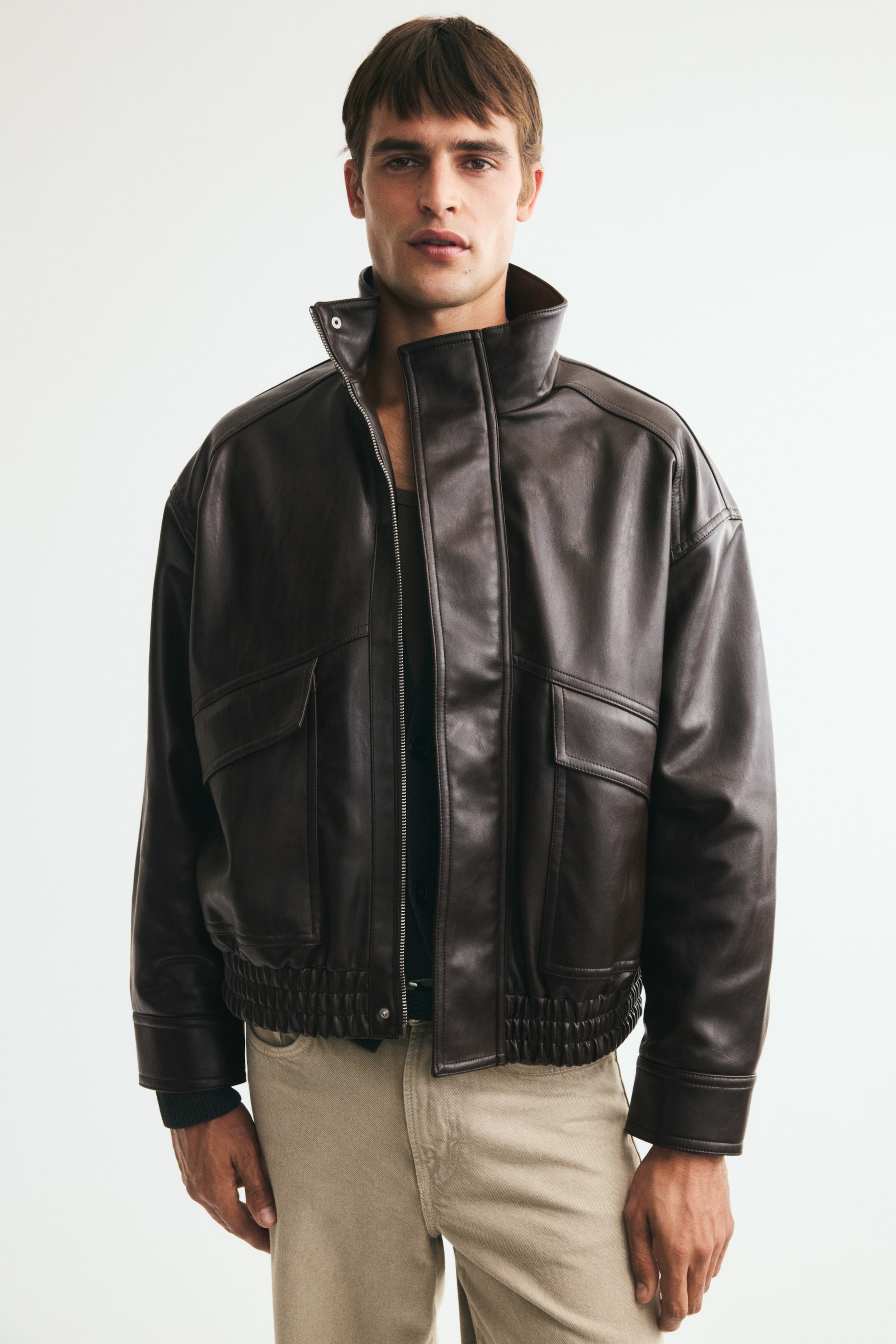 View larger image: The model is wearing a dark brown blouson jacket featuring a stand collar, a partially open front zipper with a wind flap, and dropped shoulders. The jacket features patch front pockets with flaps, press-studs at the cuffs, and smocked elastic at the hem. It is styled over a dark undershirt and light-coloured trousers.