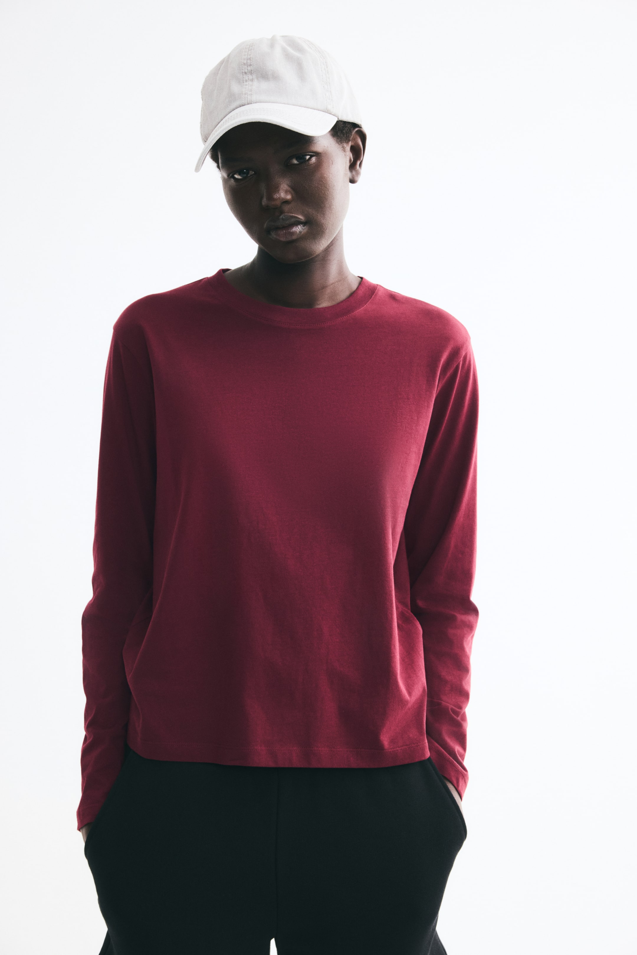 View larger image: The model is wearing a long-sleeve top in dark red cotton jersey, featuring a crew neckline, straight hem, and relaxed fit. The top is paired with black trousers and a light-colored baseball cap.