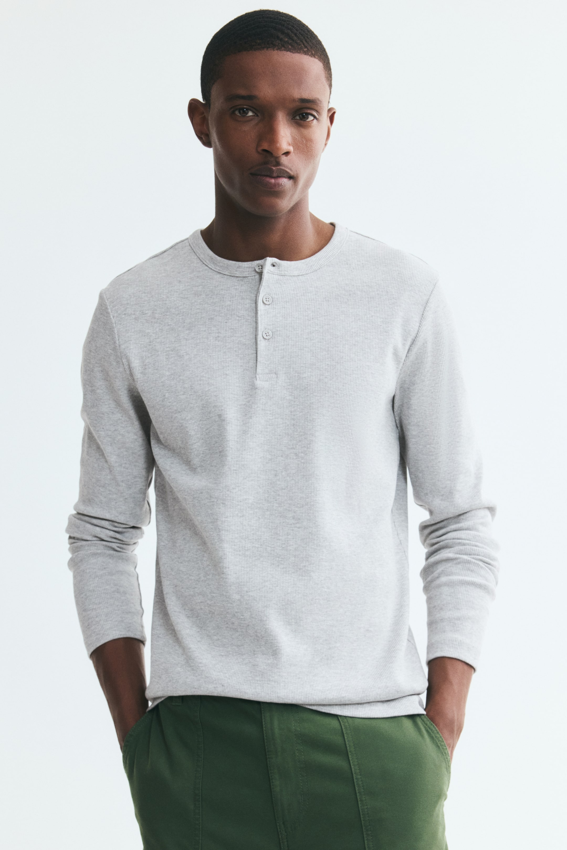 View larger image: A light grey, long-sleeve ribbed knit top featuring a round neckline and a button placket is worn by the model. The model is posing with hands in the pockets of green trousers, showcasing the slim fit of the top.