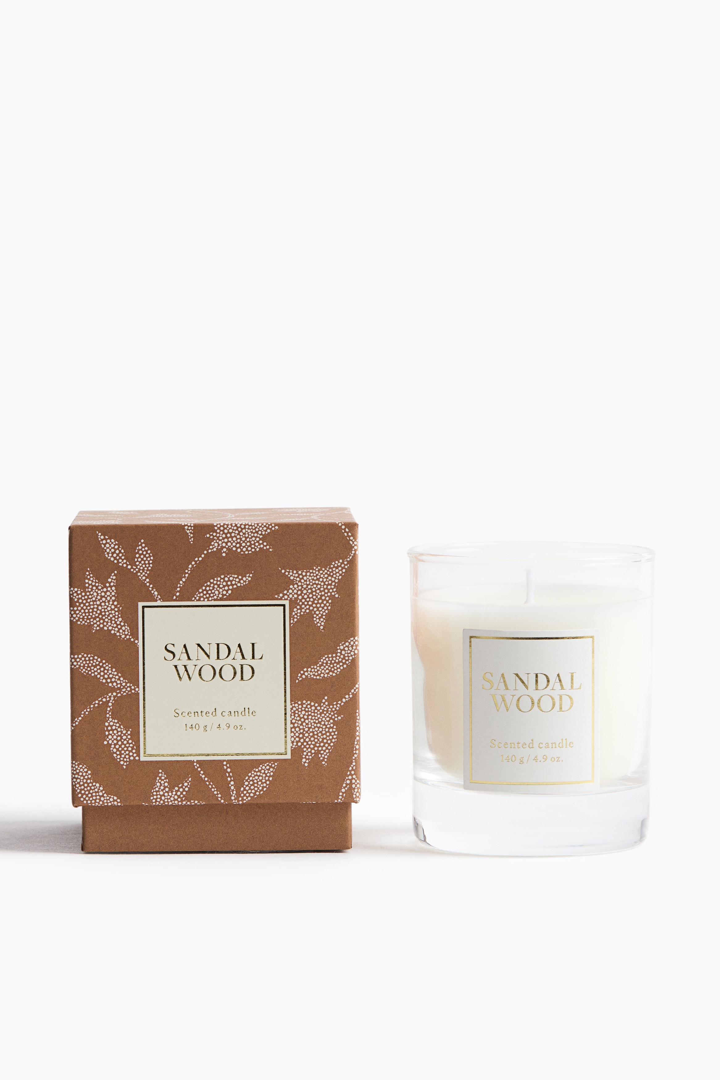 A scented candle in a clear glass holder with a thick base is resting on its side, showcasing its front-facing white label with "SANDAL WOOD" text. Surrounding it, several identical candles are displayed from a top-down perspective, showing their smooth, cream-coloured wax and central wicks.