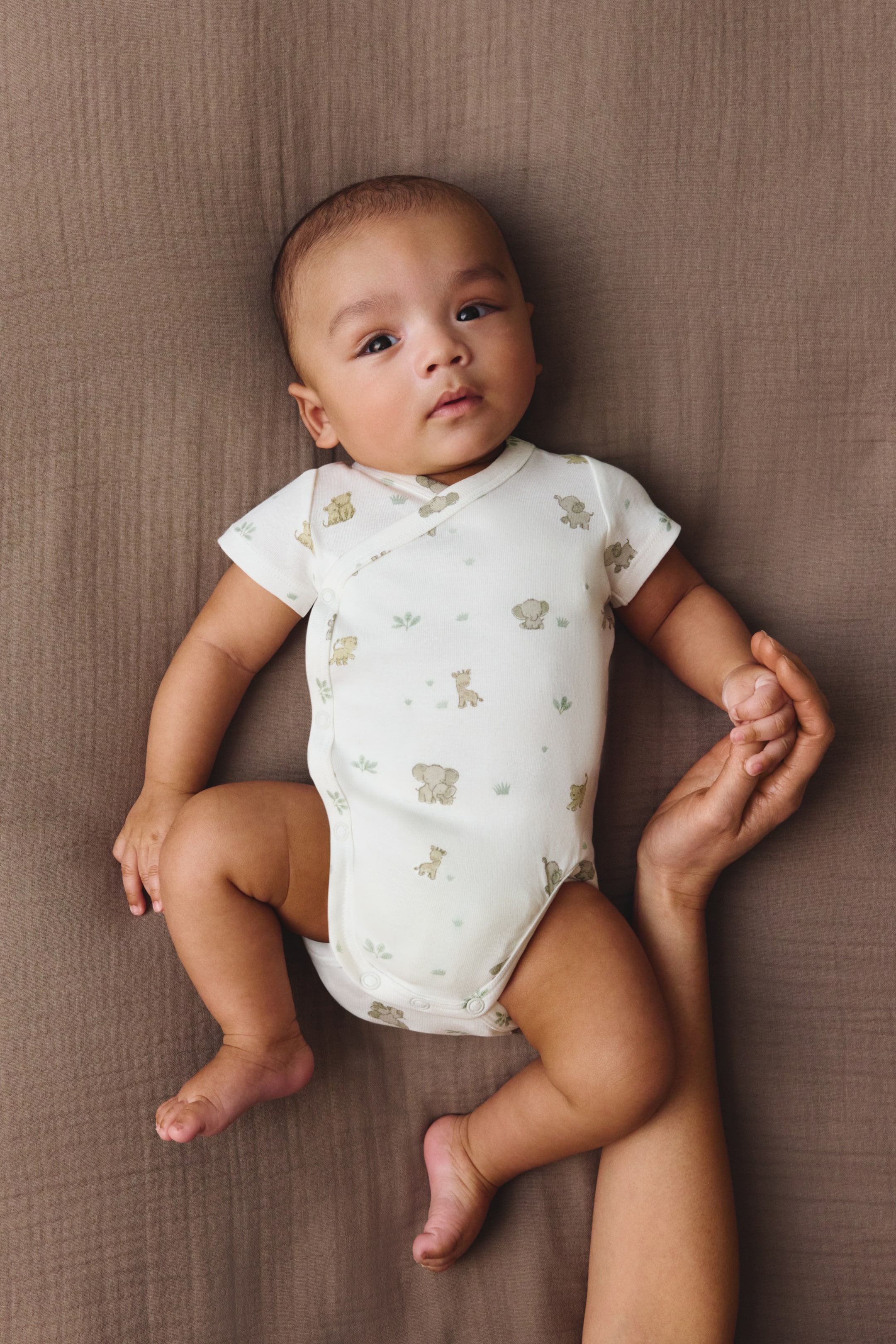 Agrandir l'image: Un body pour bébé présente une couleur blanc cassé avec un motif répétitif d'éléphants marron clair, de girafes et de feuillage vert, et est conçu avec un devant croisé de style kimono, sécurisé par des boutons-pression le long de la couture latérale et à l'entrejambe. Ce body à manches courtes est présenté en vue plongeante, mettant en valeur sa coupe.