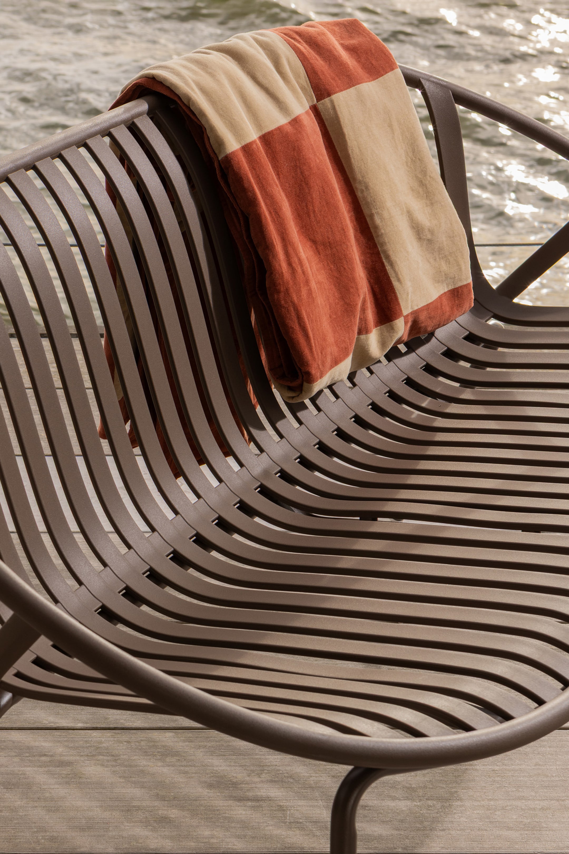 Größeres Bild anzeigen: Ein brauner Metall-Loungesessel präsentiert ein geschwungenes, lamellenartiges Design, bei dem Sitzfläche und Rückenlehne eine durchgehende, wellenförmige Oberfläche bilden, aus einem leicht erhöhten, diagonalen Winkel betrachtet. Eine gefaltete, rechteckige Decke mit abwechselnden Terrakotta- und Beigeblöcken ist über die obere Rückenlehne des Sessels drapiert.