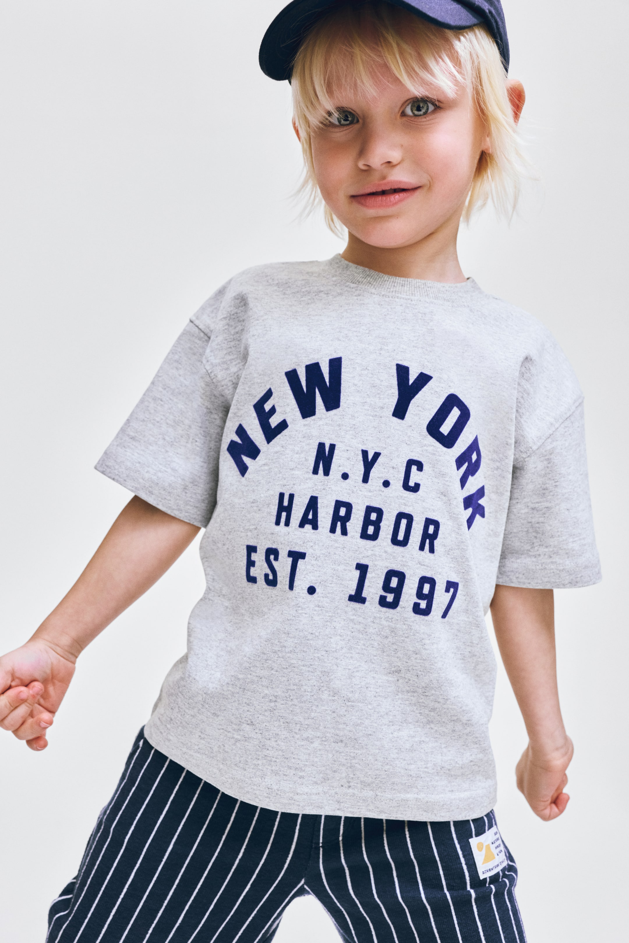 Vis større bilde: En kortermet t-skjorte med rund hals i melert grå er utstyrt med en mørkeblå grafikk på forsiden, med teksten 'NEW YORK N.Y.C HARBOR EST. 1997'. Mørkeblå bukser med tynne hvite vertikale striper bæres, med en liten gul og hvit etikett på høyre ben.