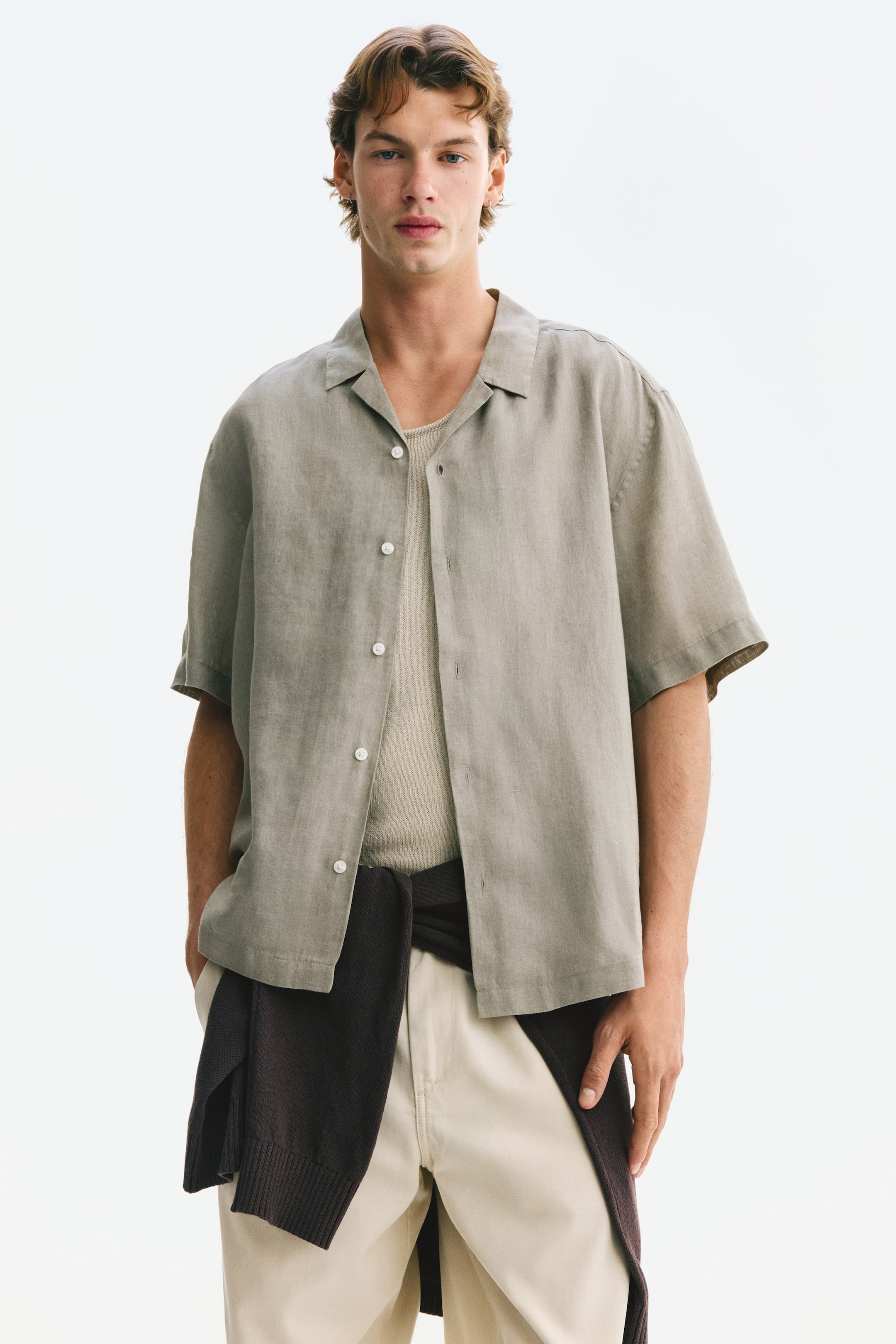 Visualizza l'immagine ingrandita: Una camicia di lino a maniche corte, con colletto alla cubana, abbottonatura francese con bottoni bianchi e orlo dritto, è indossata dalla modella. La camicia verde-grigiastro è aperta, rivelando un top di colore chiaro sotto, mentre un maglione scuro è legato intorno alla vita della modella.