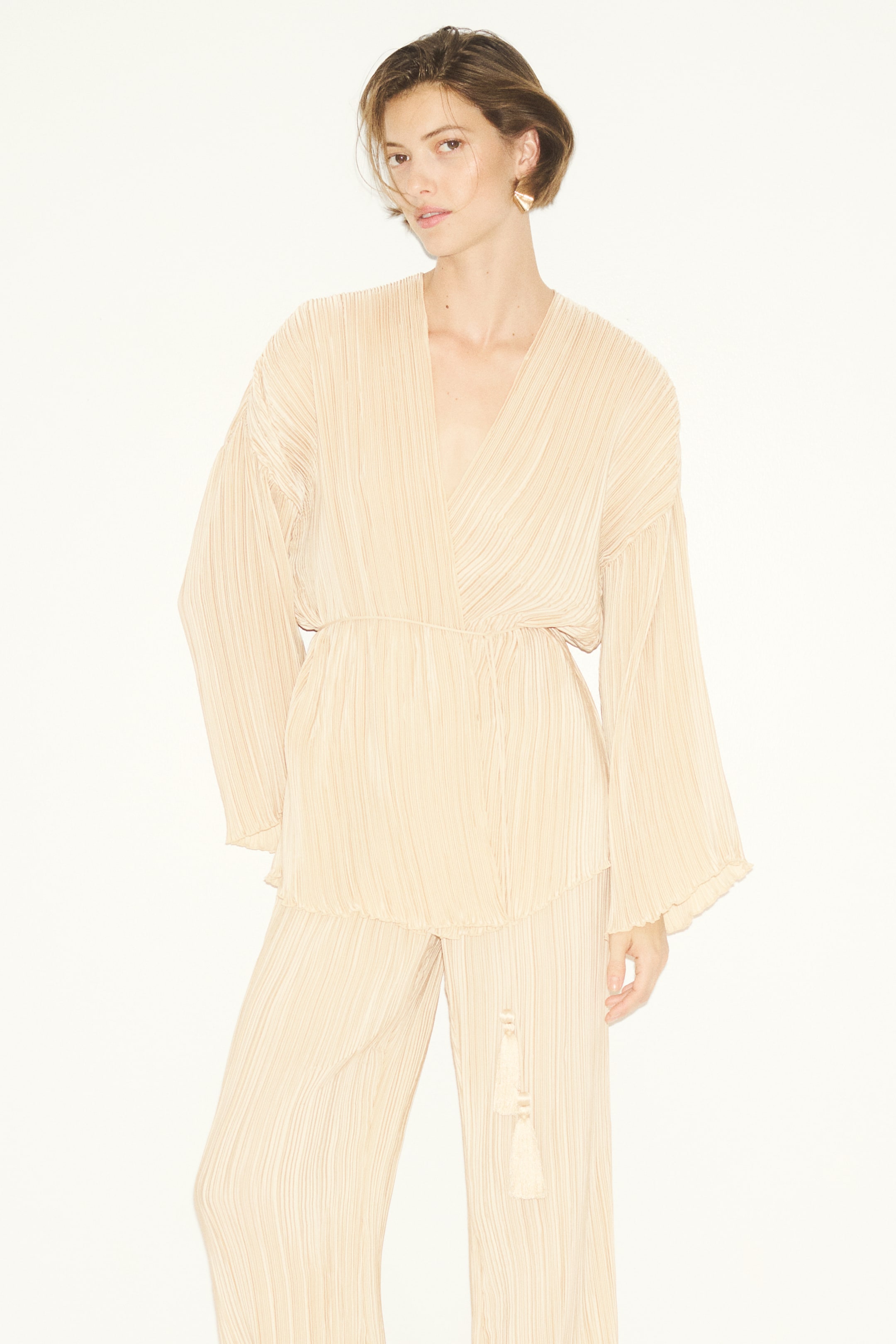 View larger image: A light beige pleated two-piece set is worn by the model, featuring a wrap-style top with a deep V-neckline and wide long sleeves. The matching wide-leg trousers feature a drawstring waist with hanging tassels.