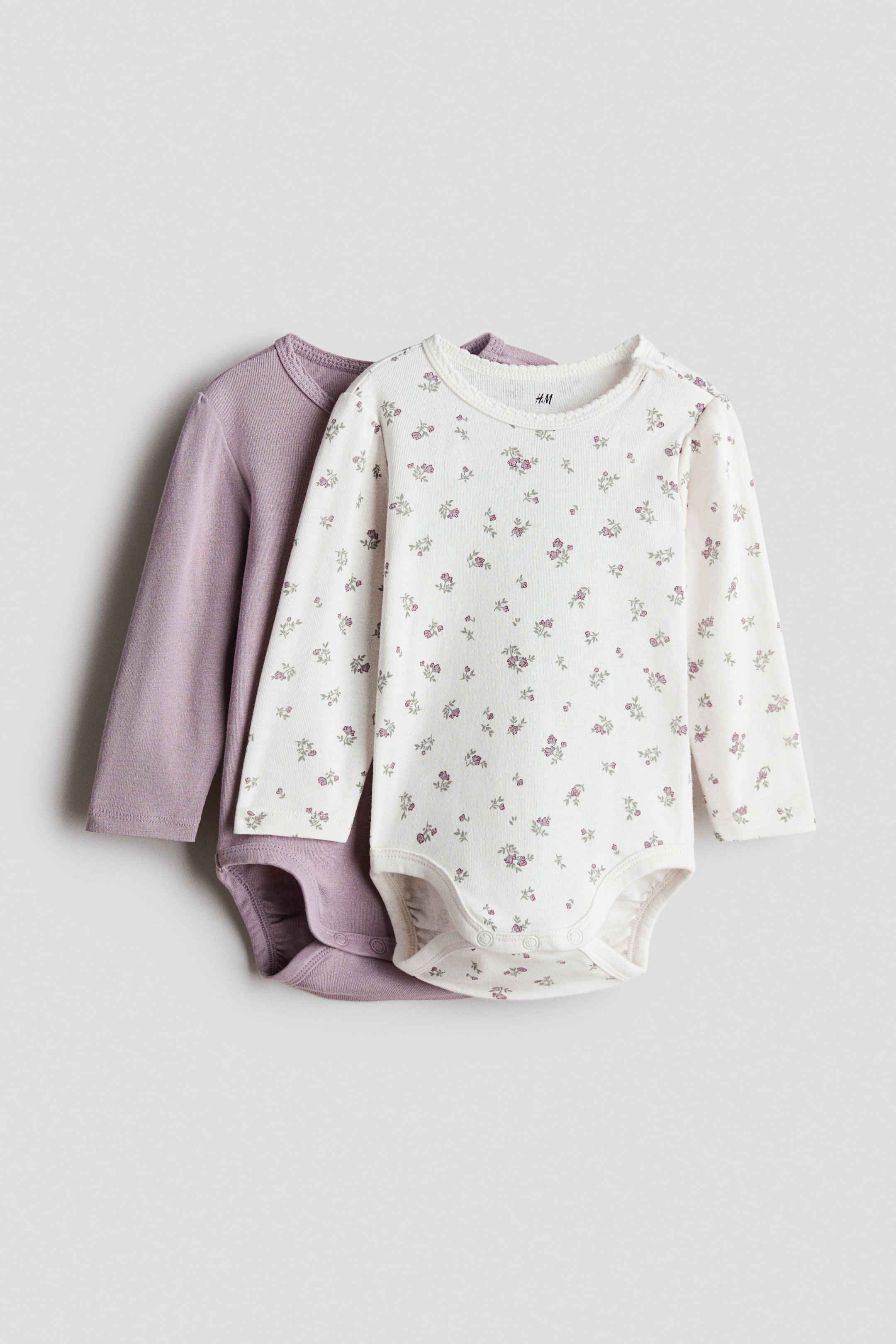 View larger image: Two long-sleeve cotton jersey bodysuits are shown in front view, one in solid light purple and the other in off-white with small purple floral patterns. Both bodysuits feature round necklines, with the off-white one displaying a delicate lace trim, and press studs are visible at the crotch and on one shoulder.