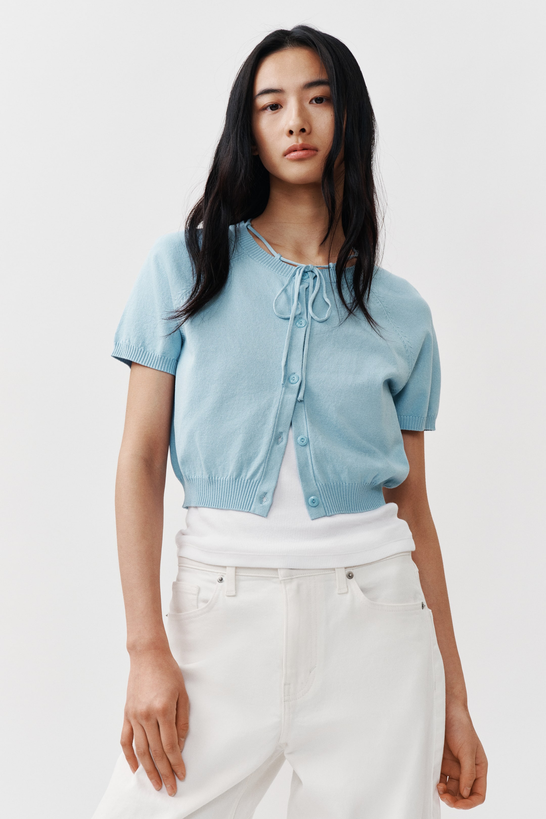View larger image: Model tersebut mengenakan kardigan pendek berwarna biru muda, dipadankan dengan tank top putih, dengan beberapa butang depan tidak dikancing. Ia menampilkan leher bulat dengan trim rib dan tali spaghetti diikat di depan, lengan raglan pendek, serta ribbing pada manset dan hem.