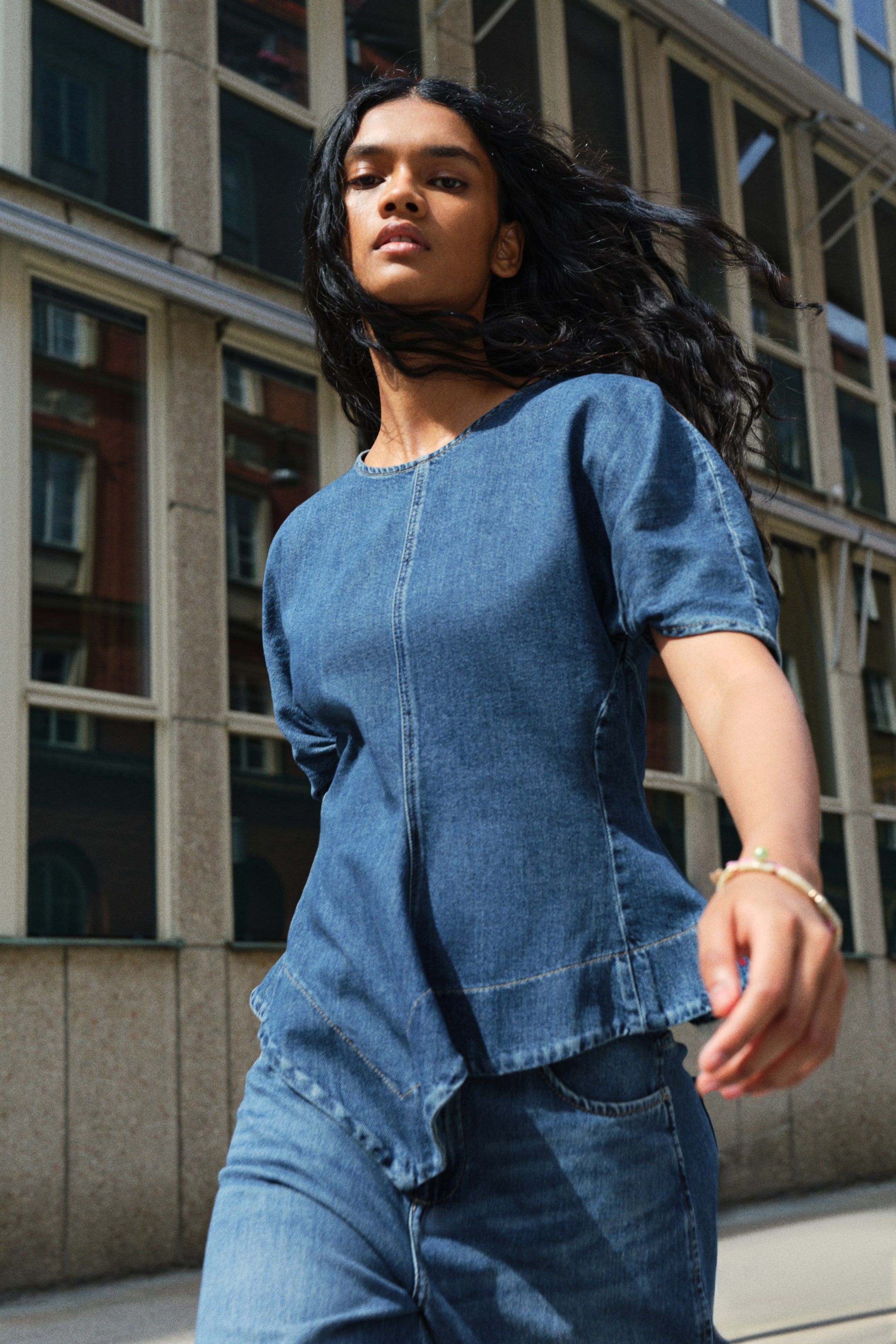 Größeres Bild ansehen: Ein blaues Oberteil aus Baumwoll-Denim mit Rundhalsausschnitt, plissierten kurzen Ärmeln und einem ausgestellten Peplum mit asymmetrischem Handkerchief-Saum wird vom Model mit passender Denim-Jeans getragen.