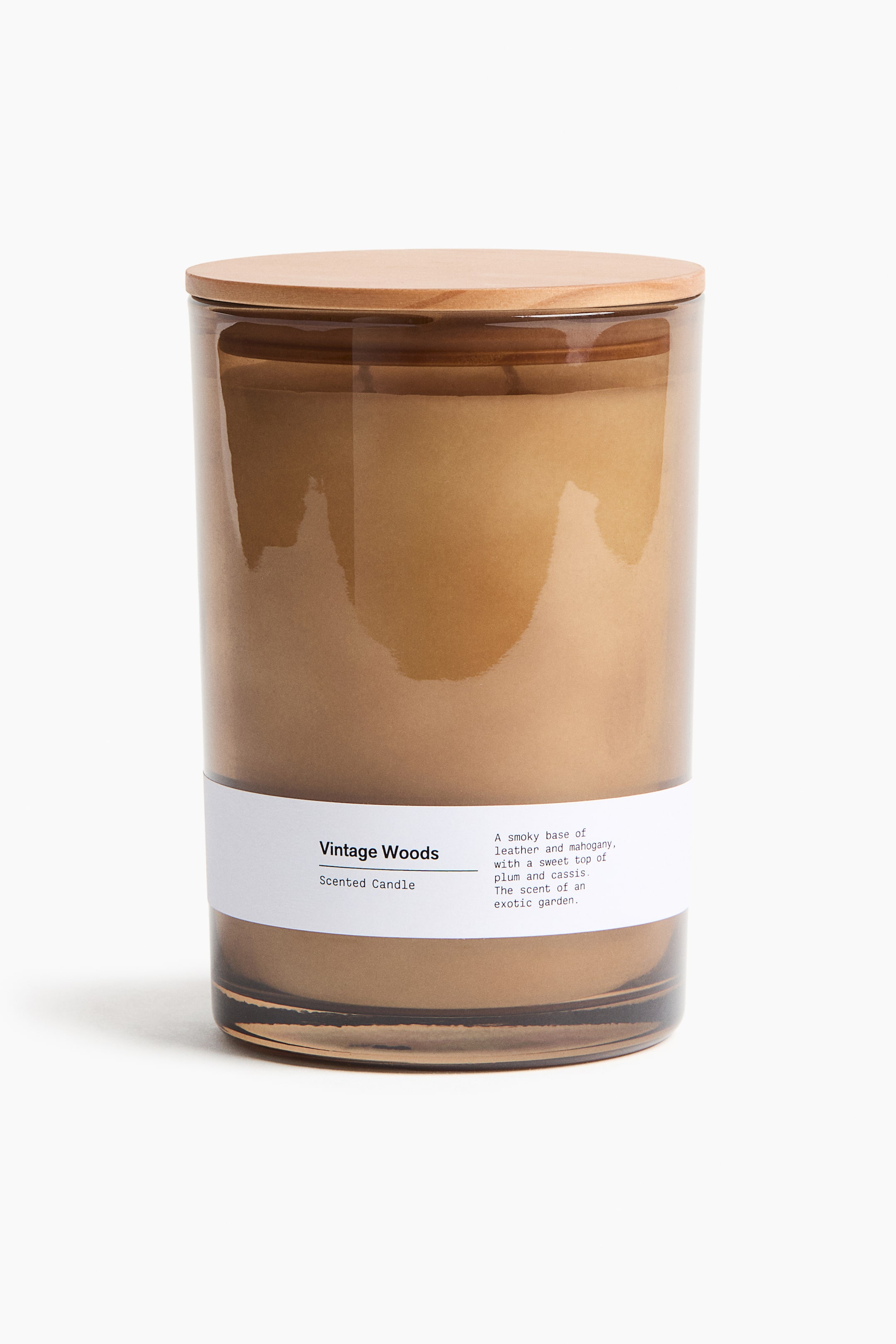 View larger image: A scented candle is standing front-facing in a cylindrical amber glass holder, topped with a light-colored pinewood lid. A white rectangular label is wrapping around the lower half, displaying "Vintage Woods Scented Candle" and a scent description.