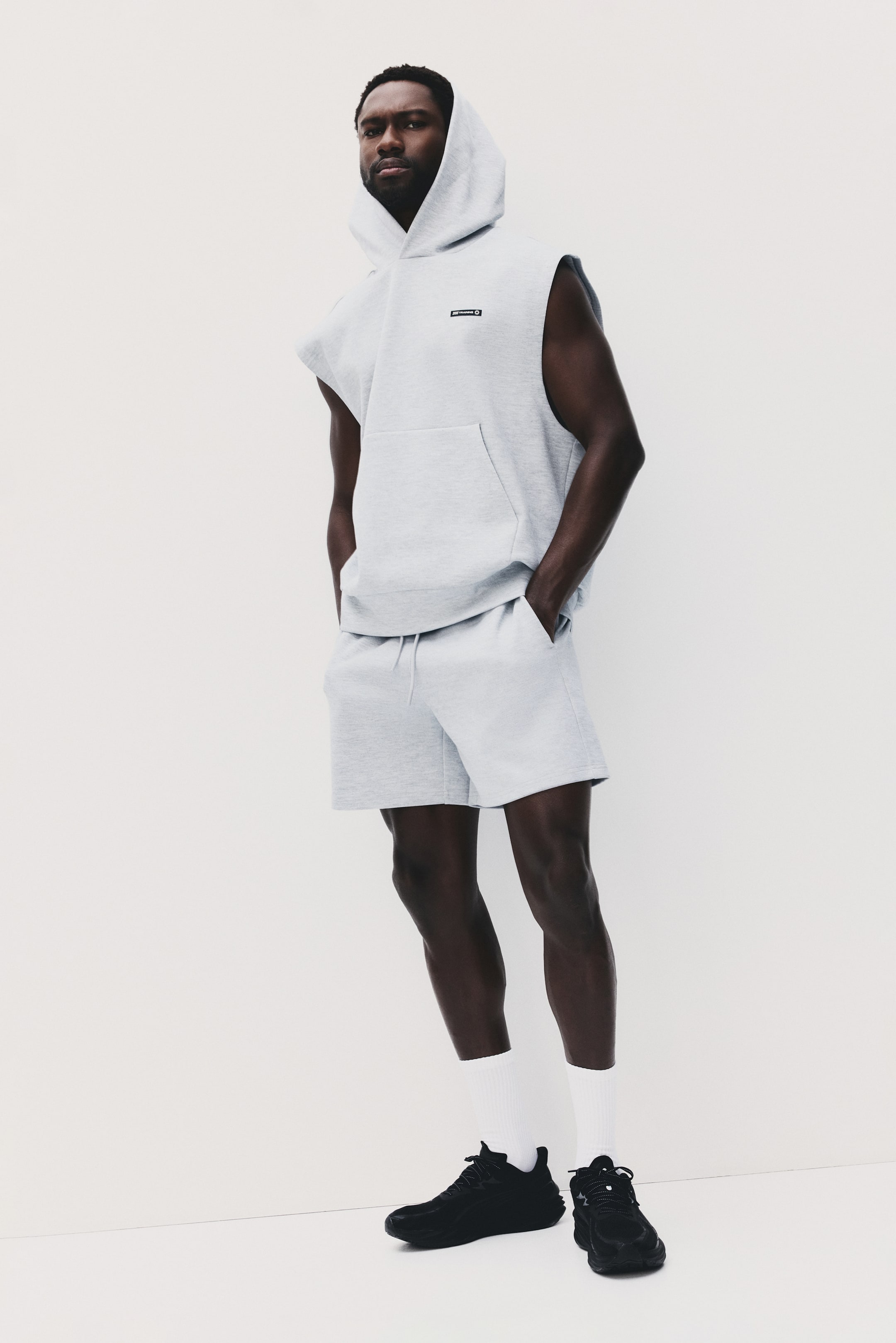 View larger image: Presenting a light grey sleeveless hoodie and matching shorts set, positioned slightly to the left. The hoodie comes with a drawstring hood, a front kangaroo pocket, and a rectangular logo patch. The shorts feature a drawstring waist and side pockets.