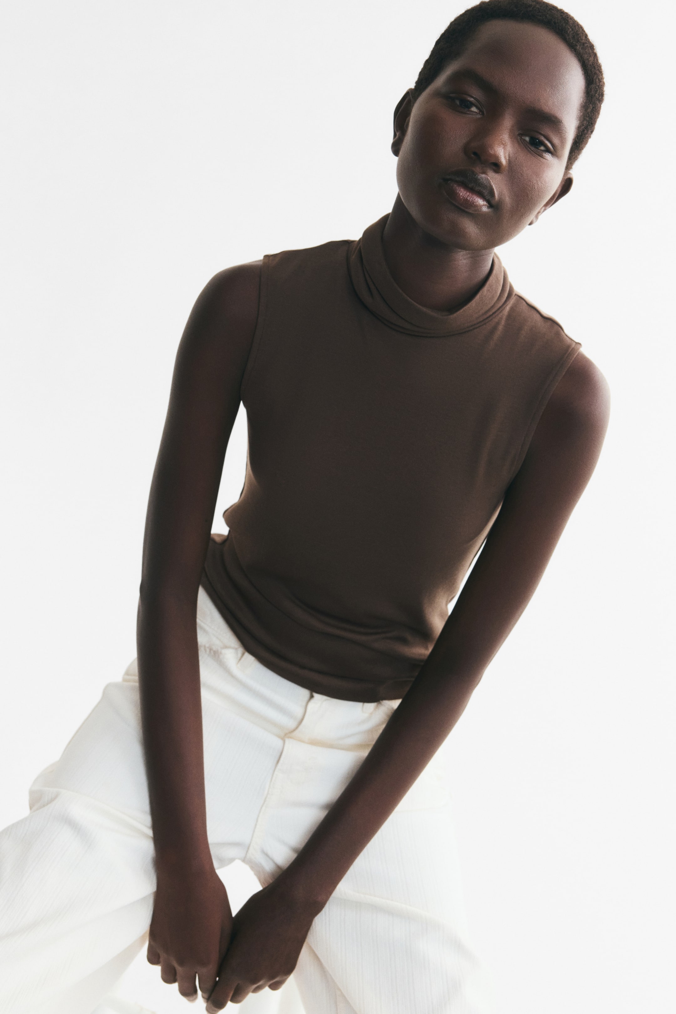 View larger image: A brown sleeveless top featuring a draped high neckline is being worn by the model and styled with light-coloured wide-leg trousers.