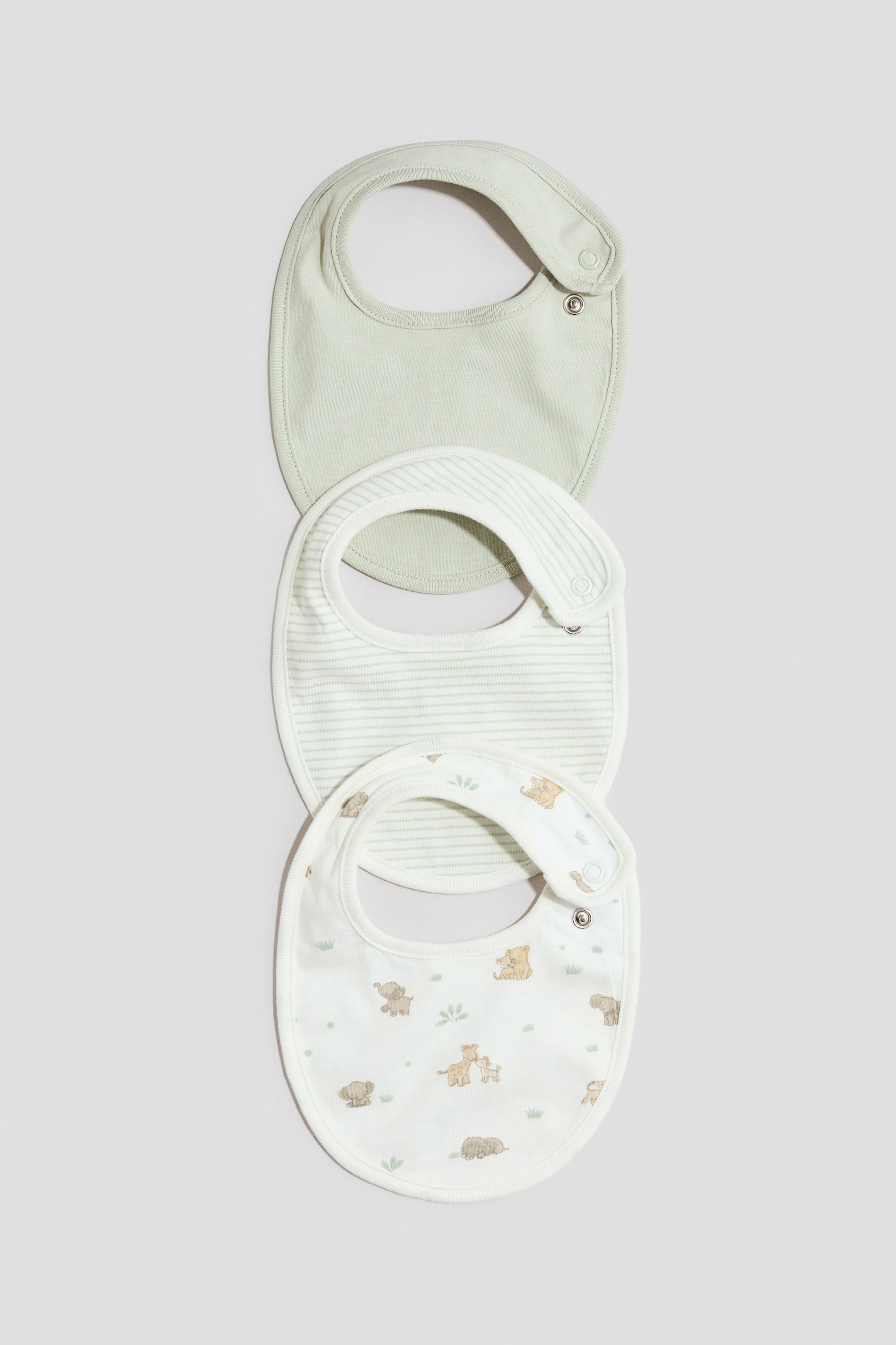 Visualizza immagine più grande: Tre bavaglini in morbido jersey di cotone sono esposti FRONTE, impilati verticalmente. Il bavaglino superiore è di un verde chiaro solido, il bavaglino centrale è bianco con strisce orizzontali verde chiaro e il bavaglino inferiore è bianco con un animale cartone marrone e stampa di piante verdi, ciascuno dotato di una chiusura a pressione regolabile su un lato.