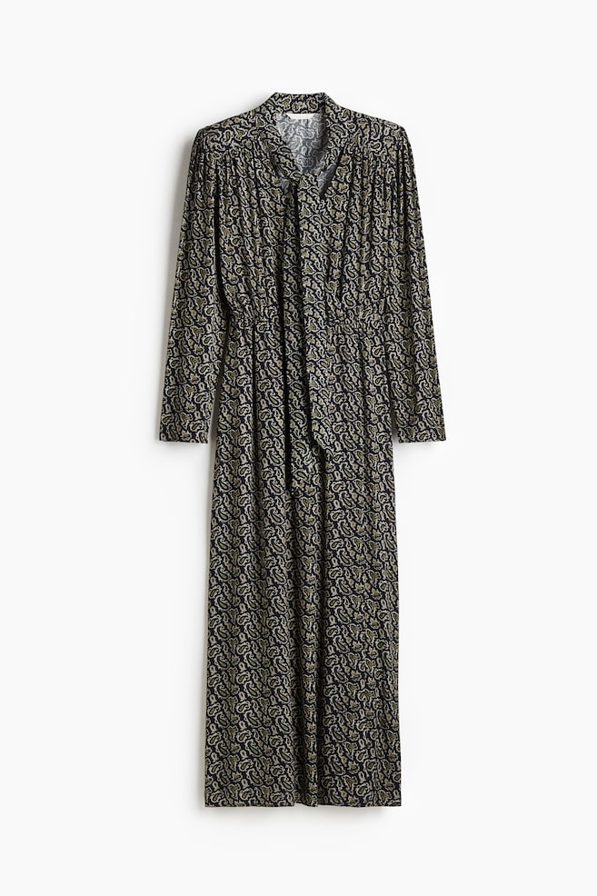 Ein langärmliges Maxikleid wird von vorne gezeigt, mit einem schwarzen Stoff und einem hellen Paisleymuster. Das Kleid ist mit einem Stehkragen gestaltet, der zu einem V-Ausschnitt mit Bindedetails führt, einer elastischen Taille und einem fließenden, gerafften Rock. - 1