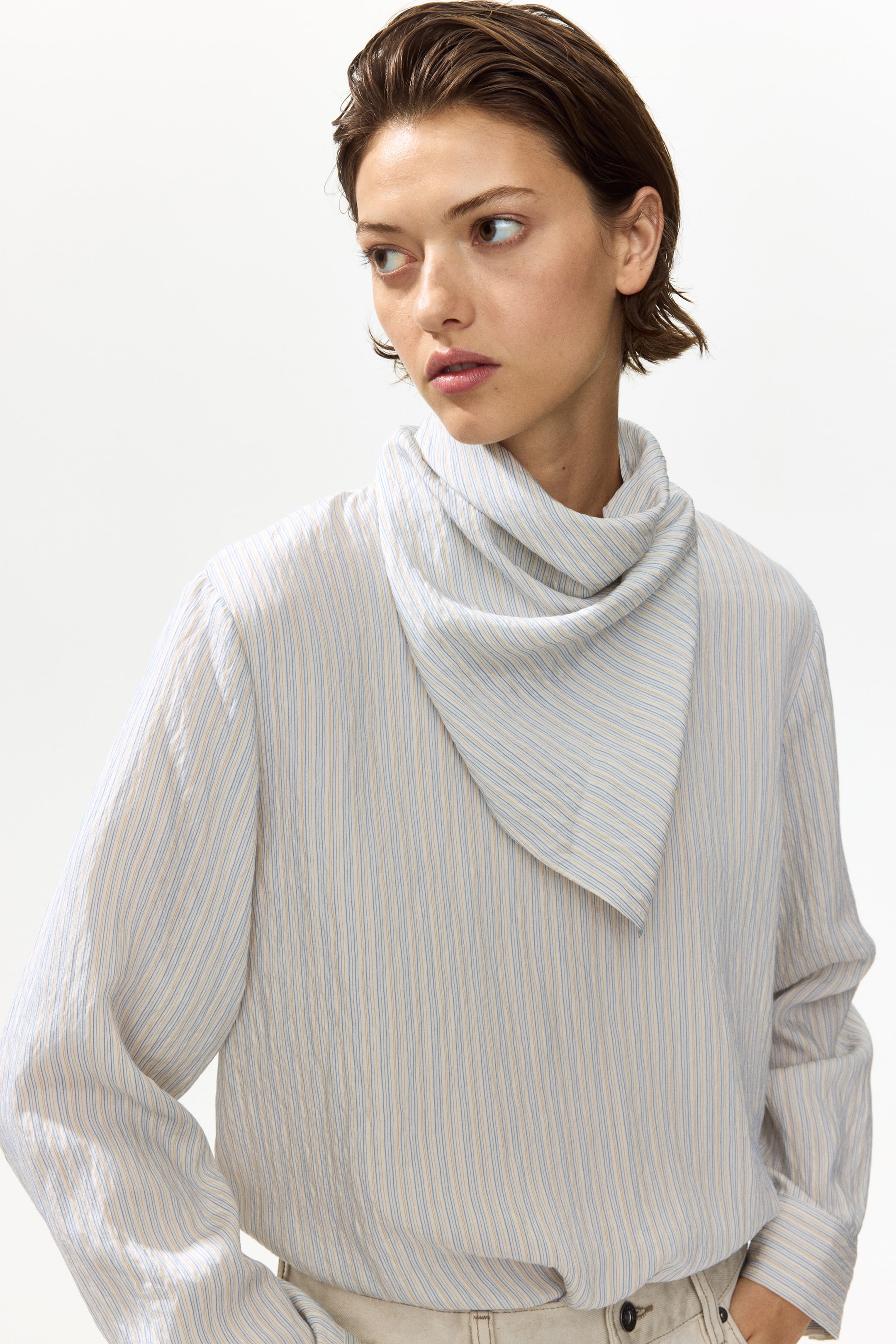 Grotere afbeelding bekijken: Een blouse met textuur, met dunne blauwe en beige verticale strepen en lange mouwen, wordt gedragen door het model. Een bijpassende gestreepte driehoekige sjaal is om de hals van het model gedrapeerd.