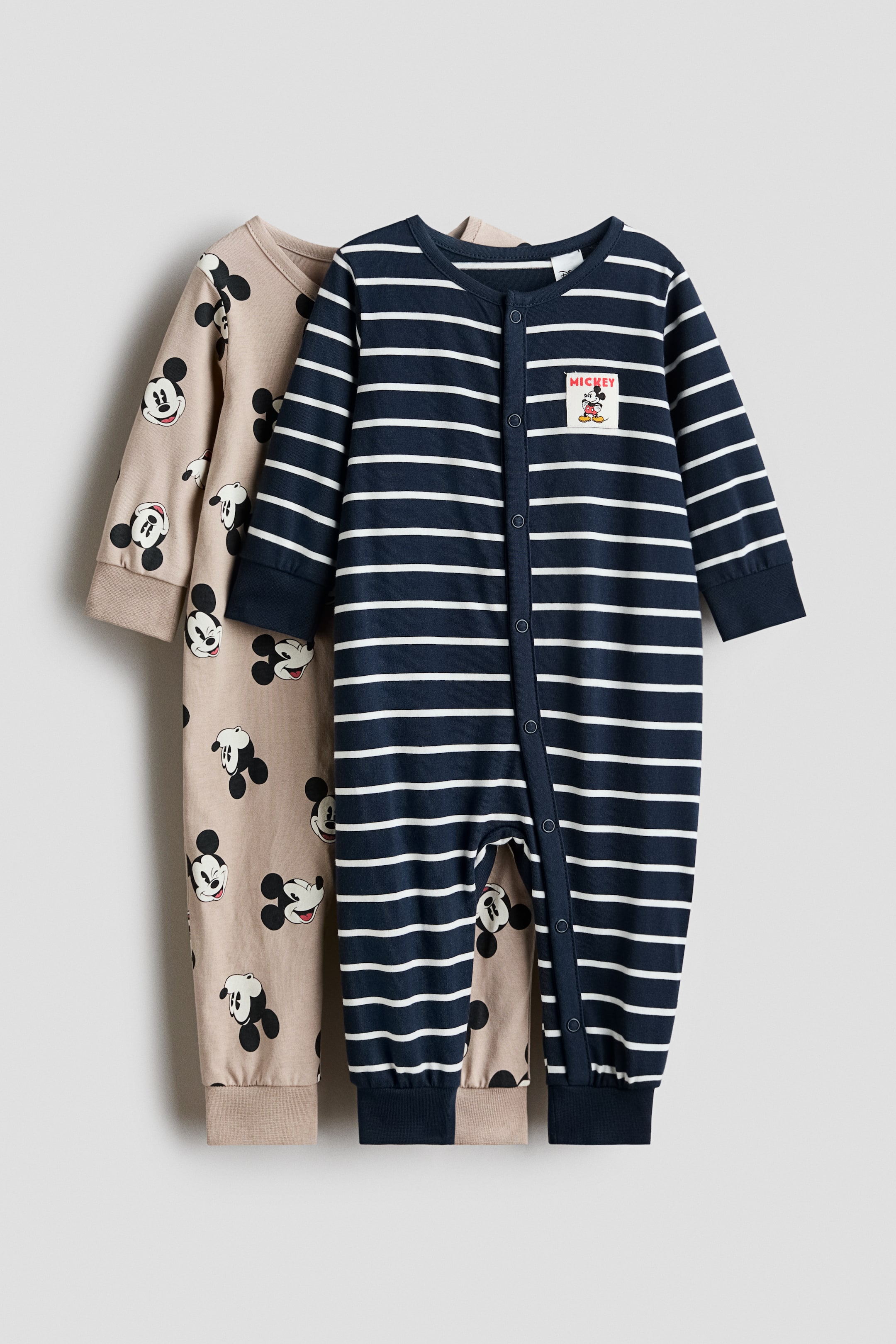 View larger image: Two cotton jersey sleepsuits are displayed with their front designs visible: one features a navy and white striped pattern with a small chest patch, while the other showcases an all-over Mickey Mouse print on a beige background. Both sleepsuits feature press-stud fastenings running down the front and along one leg, finished with wide ribbed cuffs at the sleeves and hems.