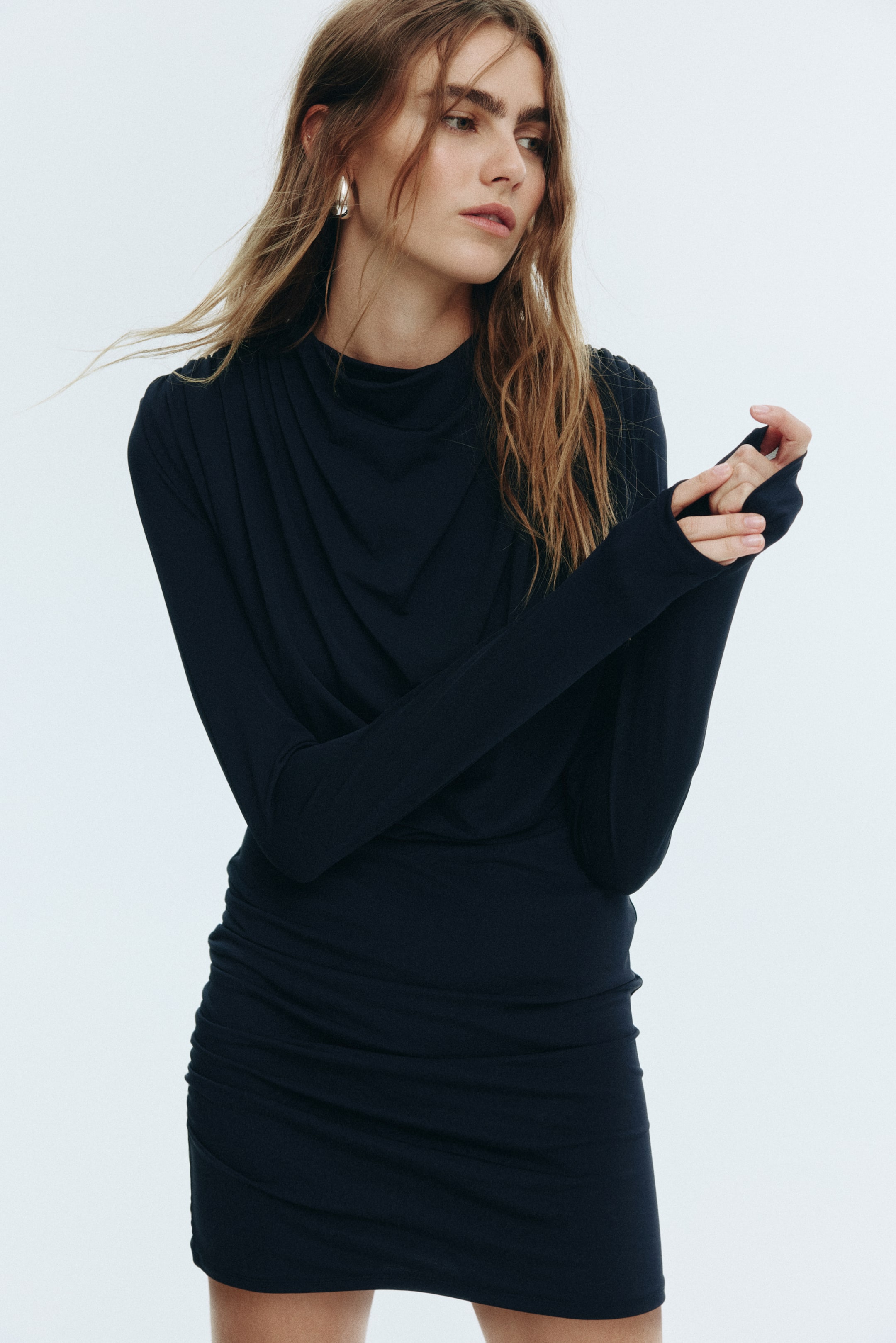 View larger image: A black long-sleeved mini-dress, seen from a front-right angle, is featuring a draped high neckline, thumbhole cuffs, and ruching details on the side of the skirt.