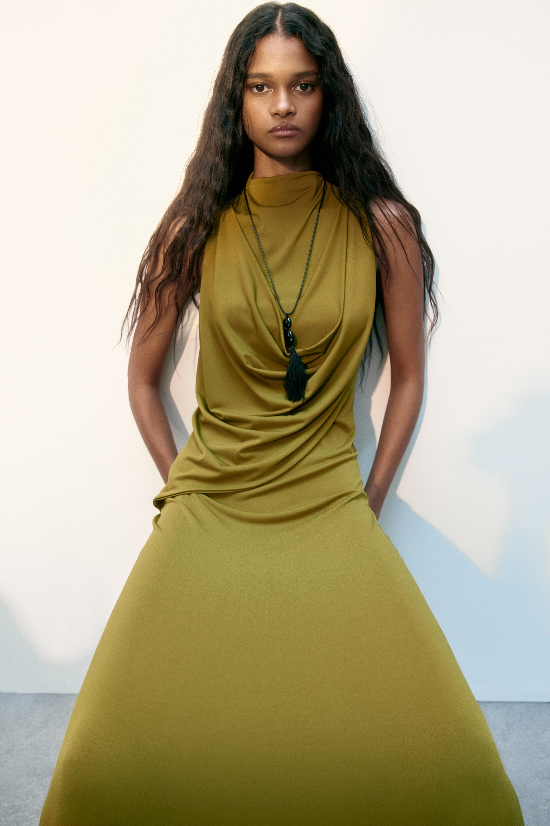 View larger image: An olive green sleeveless maxi dress is crafted with a high neck that gracefully drapes into a cowl neck, complemented by a flowing, floor-length skirt. A long black necklace featuring two oval beads and a tassel elegantly rests on the cowl neck.