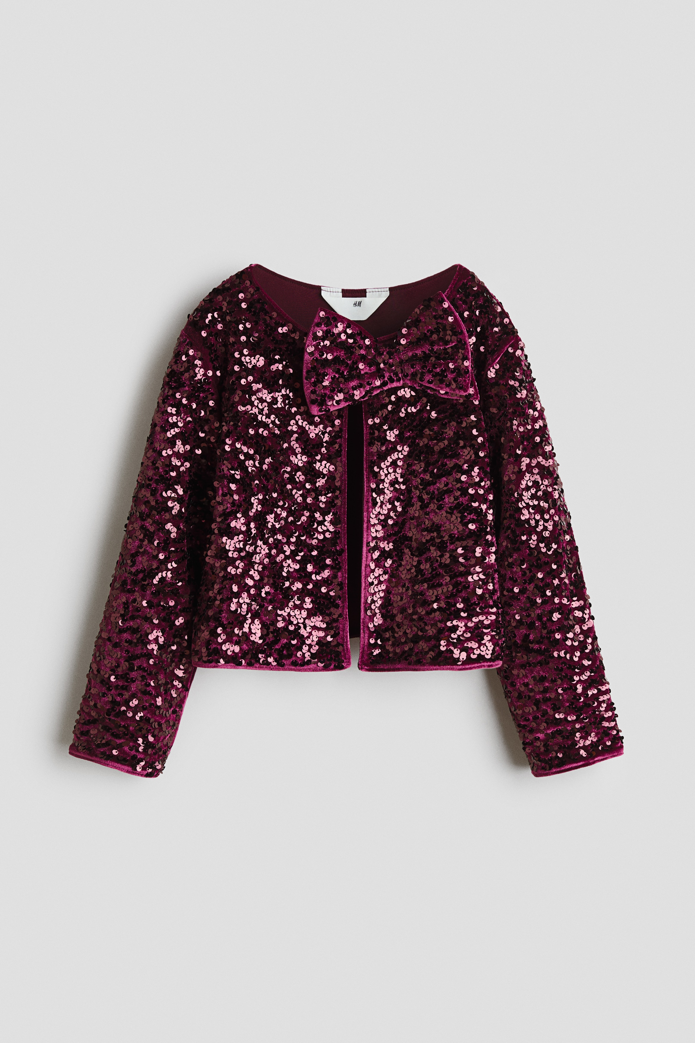 A dark pink, sequin-covered velour jacket is shown from the front, featuring a round neckline and a large decorative bow at the top closure. The jacket is worn open over a white top, highlighting its shimmering texture and structured design.