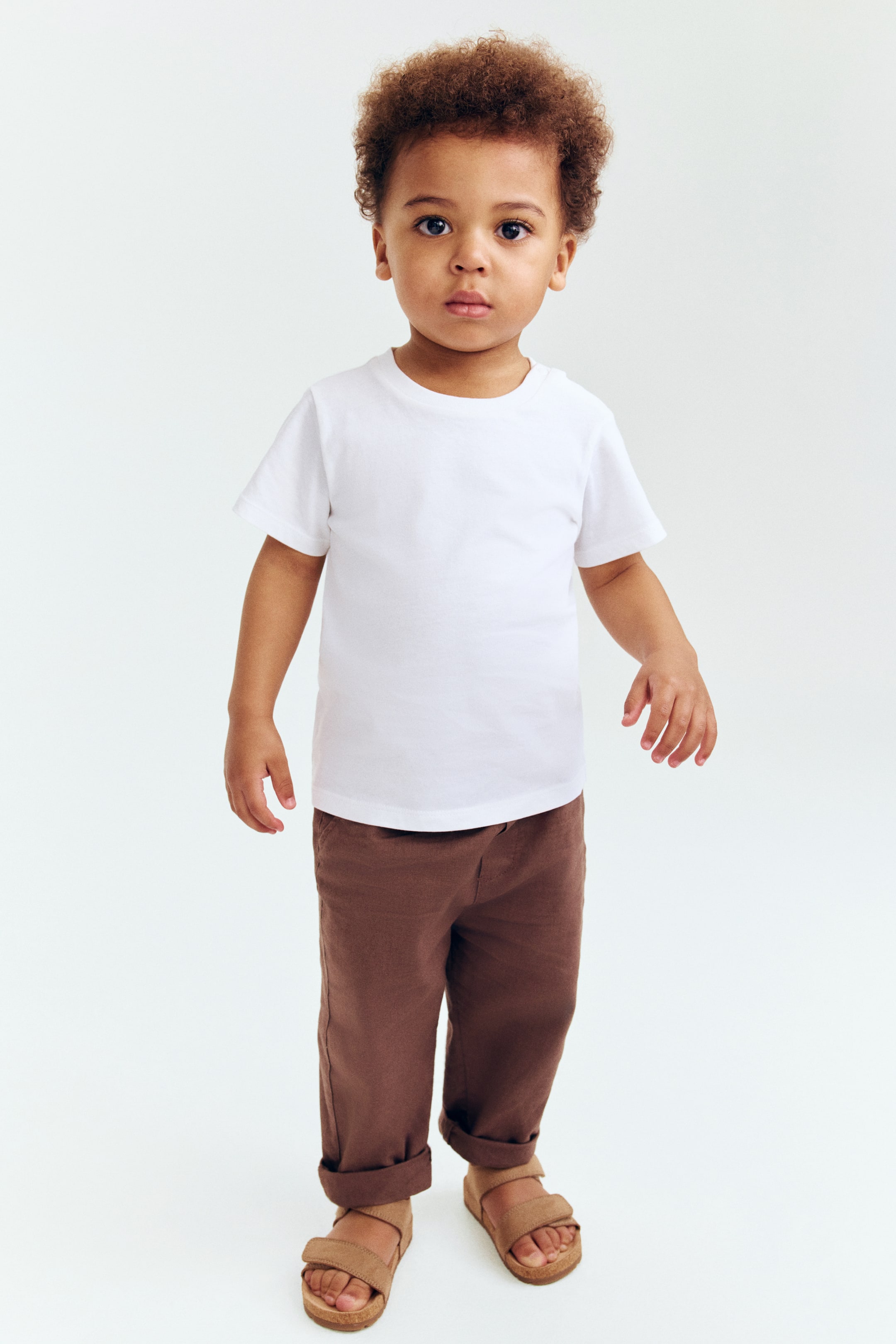 View larger image: A plain white short-sleeve t-shirt with a crew neck is worn, paired with brown loose-fit pants featuring cuffed hems and tan two-strap sandals with cork-style soles.