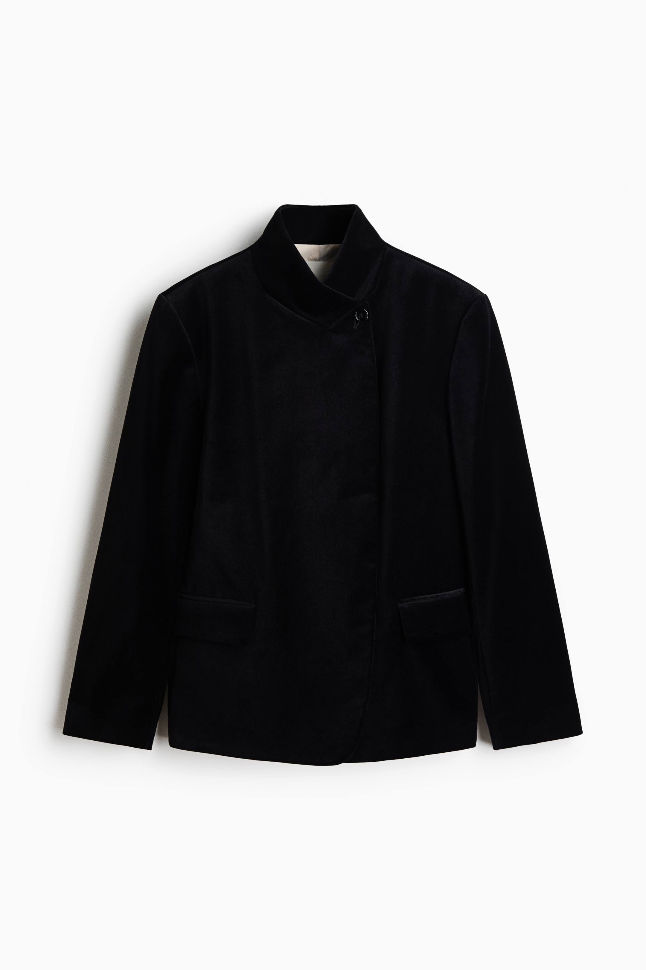 View larger image: A black velvet jacket is hanging front-facing, featuring an asymmetrical closure with a single button at the high stand-up collar. The jacket displays structured shoulders, long straight sleeves, and two lower front flap pockets.