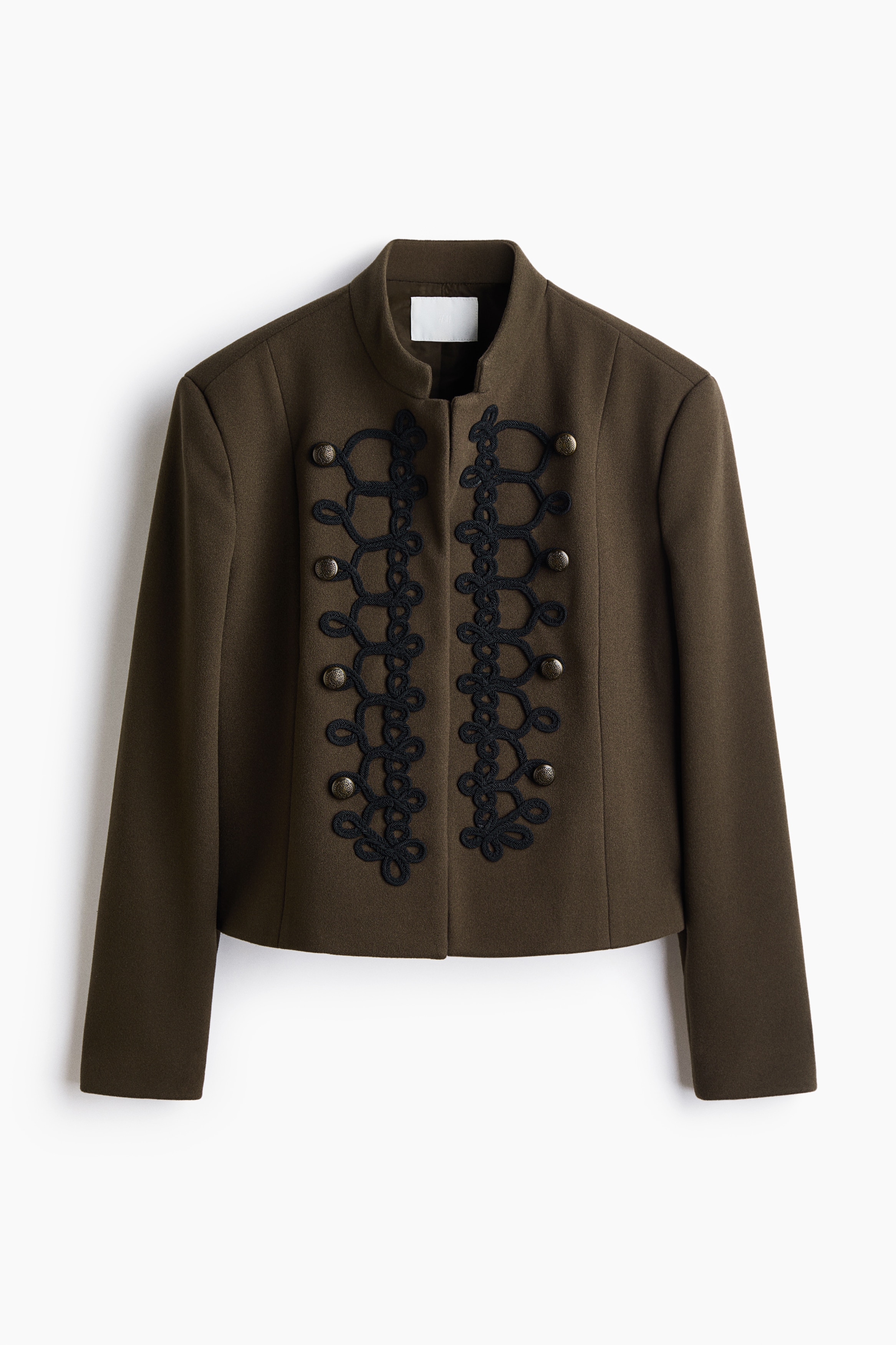A short olive-green jacket in a felted fabric is worn by the model over a black top and black leather trousers. The jacket features a band collar, an open front, decorative black braided trims, and metallic spherical buttons.