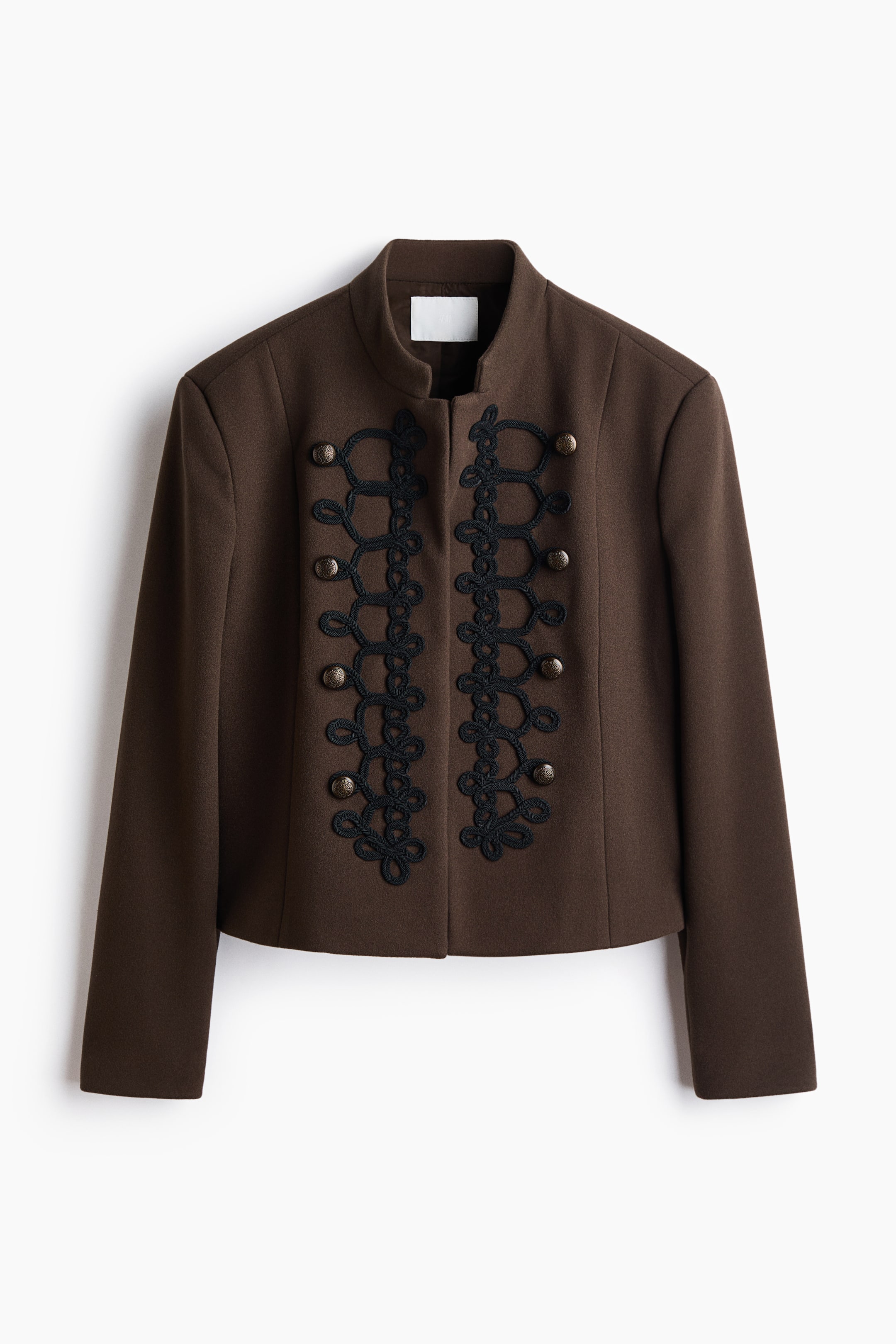 Ampliar la imagen: Una chaqueta corta estilo militar en color café está de frente, con cuello alto y mangas largas. Un intrincado bordado de trenzado negro tipo frogging recorre ambos lados de la abertura frontal, acentuado por cuatro botones metálicos tipo domo en cada lado.