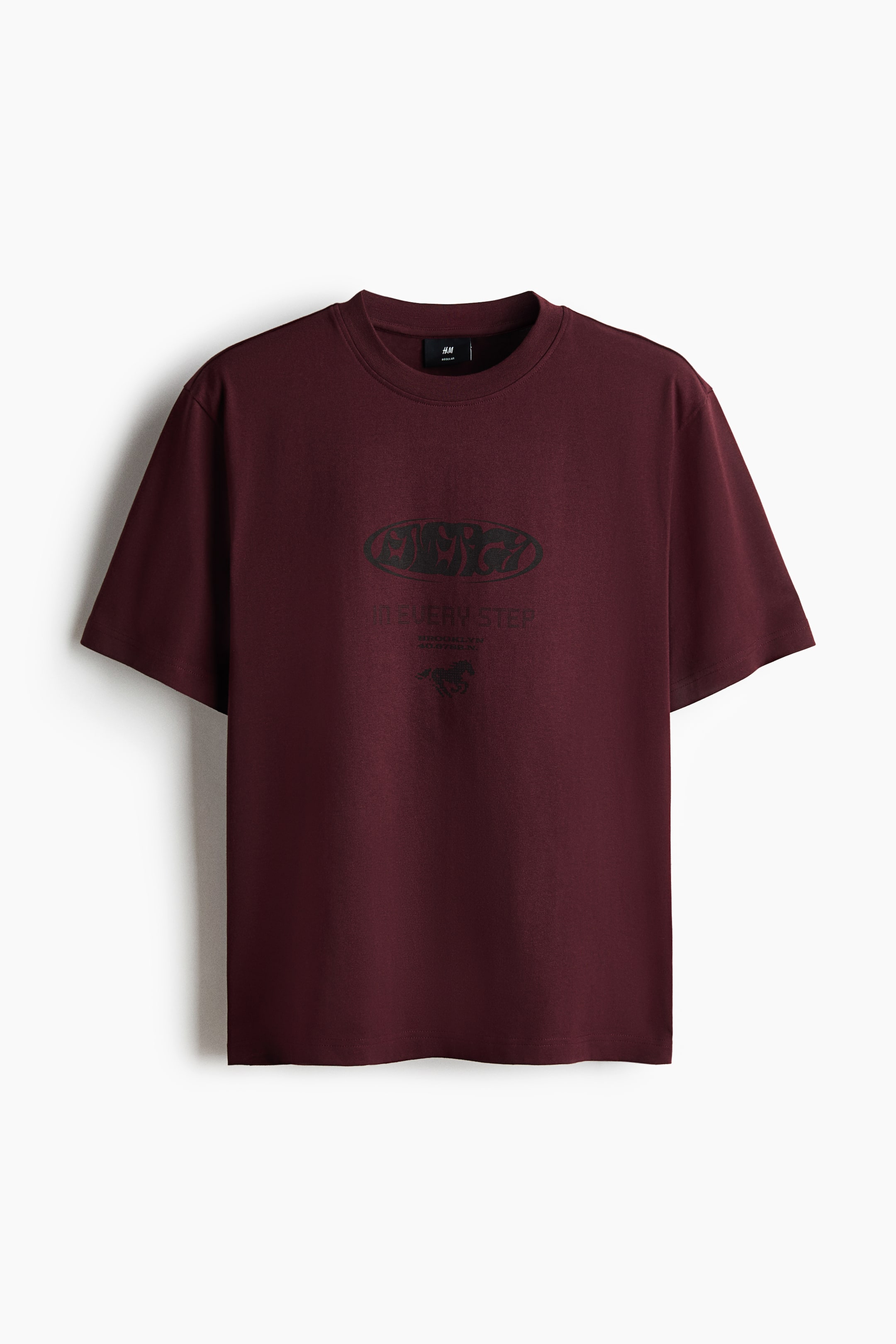 View larger image: A maroon T-shirt is shown from the front, featuring a ribbed crew neck and short sleeves. A dark graphic print is placed at the centre of the chest, displaying an oval design with abstract lettering, the text "IN EVERY STEP" below it, and a small emblem at the bottom.