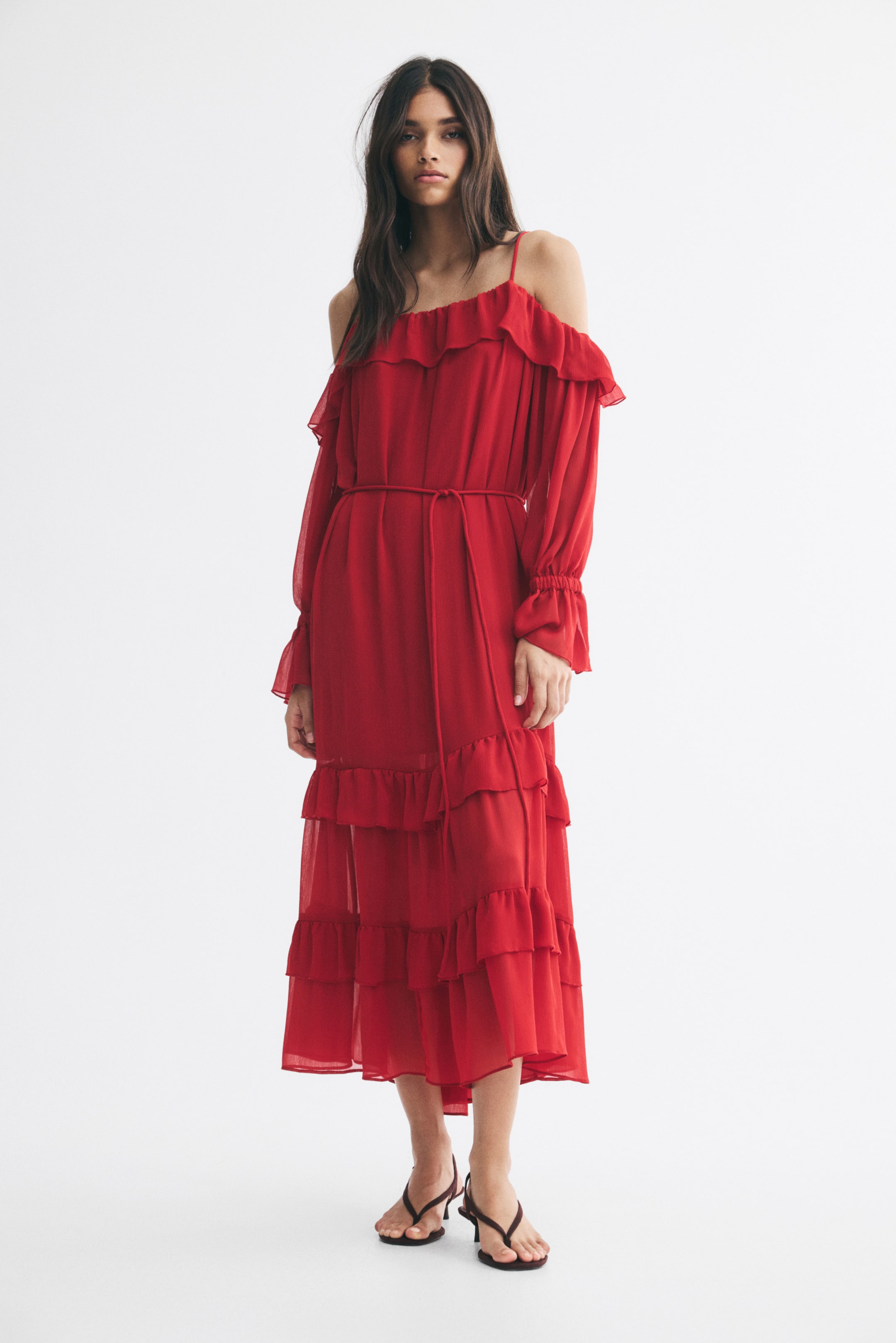 View larger image: The model is wearing a red off-the-shoulder dress with frill trims. The midi-length dress features thin shoulder straps, a ruffled neckline, long sleeves with elasticated cuffs, a self-tie waist belt, and a tiered ruffled skirt.