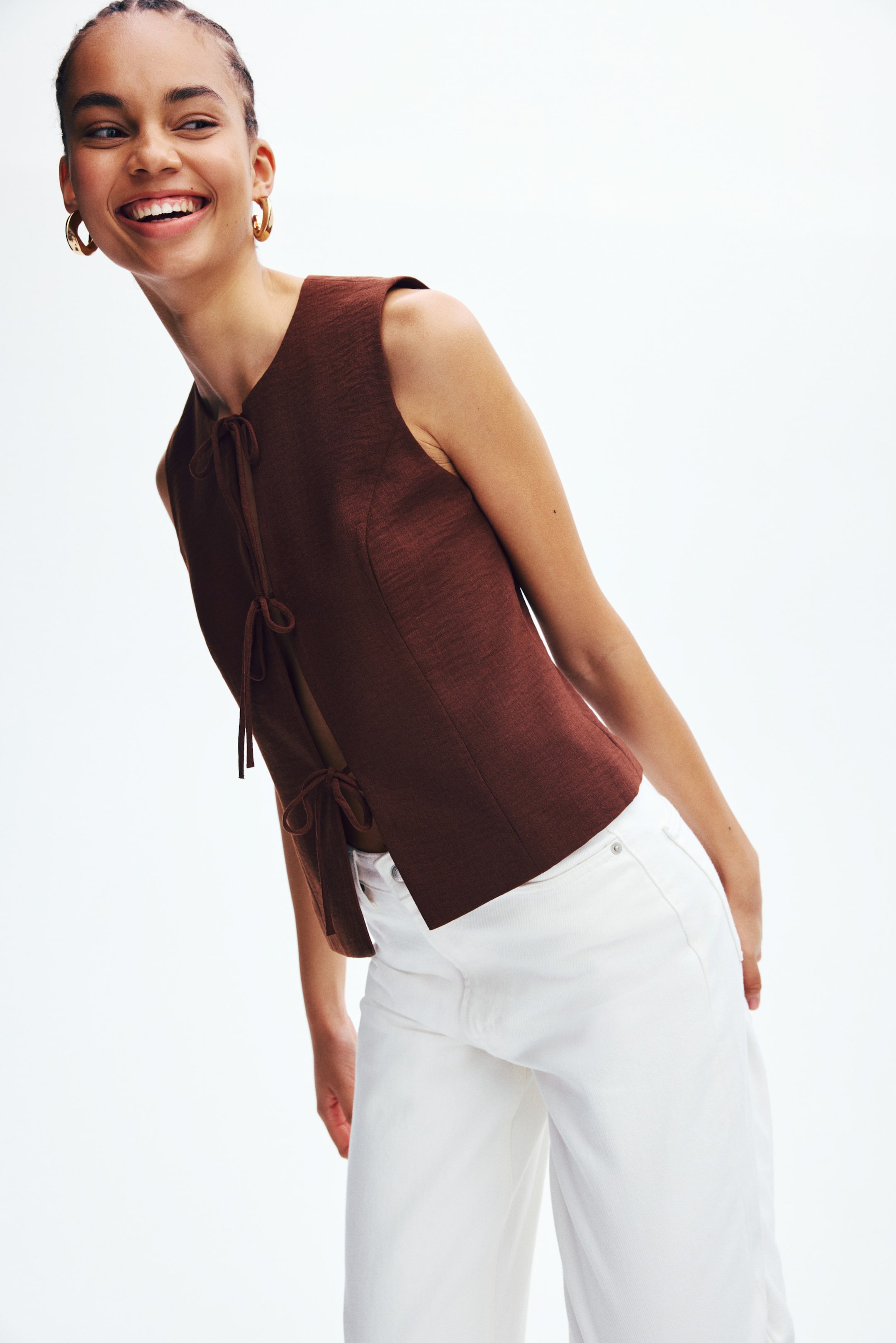 View larger image: A textured brown sleeveless top showcases a V-neckline and multiple ties along the front and back, offering an open design. The top is tailored at the waist and is paired with white high-waisted trousers.