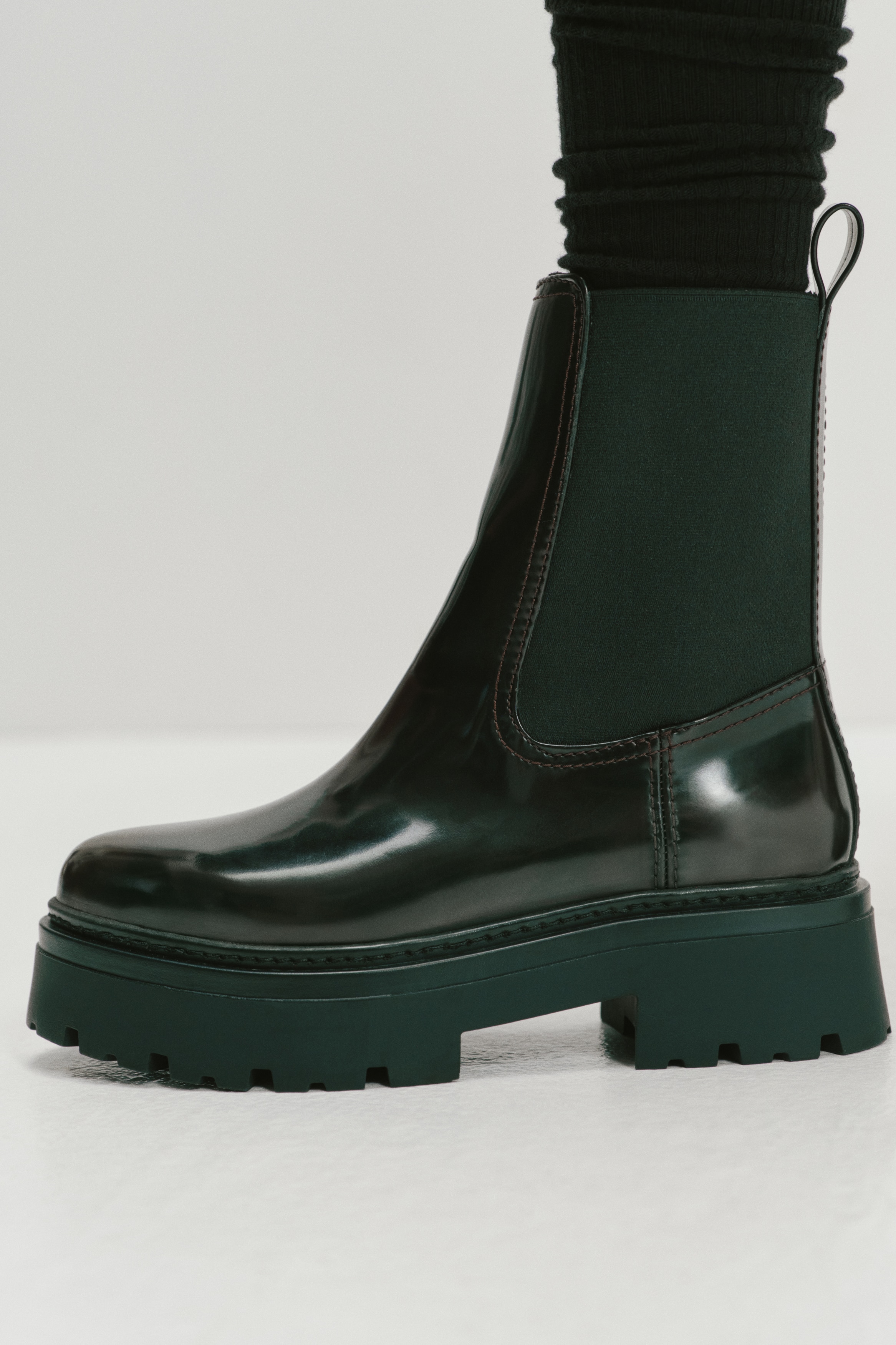 Ein dunkelburgunderfarbener Chelsea-Boot aus Leder ist in der Seitenansicht zu sehen und präsentiert sein glattes, poliertes Obermaterial sowie einen schwarzen elastischen Seiteneinsatz. Der Boot verfügt über eine dicke, schwarze Profilsohle aus Gummi und eine Zuglasche am hinteren Schaftrand.