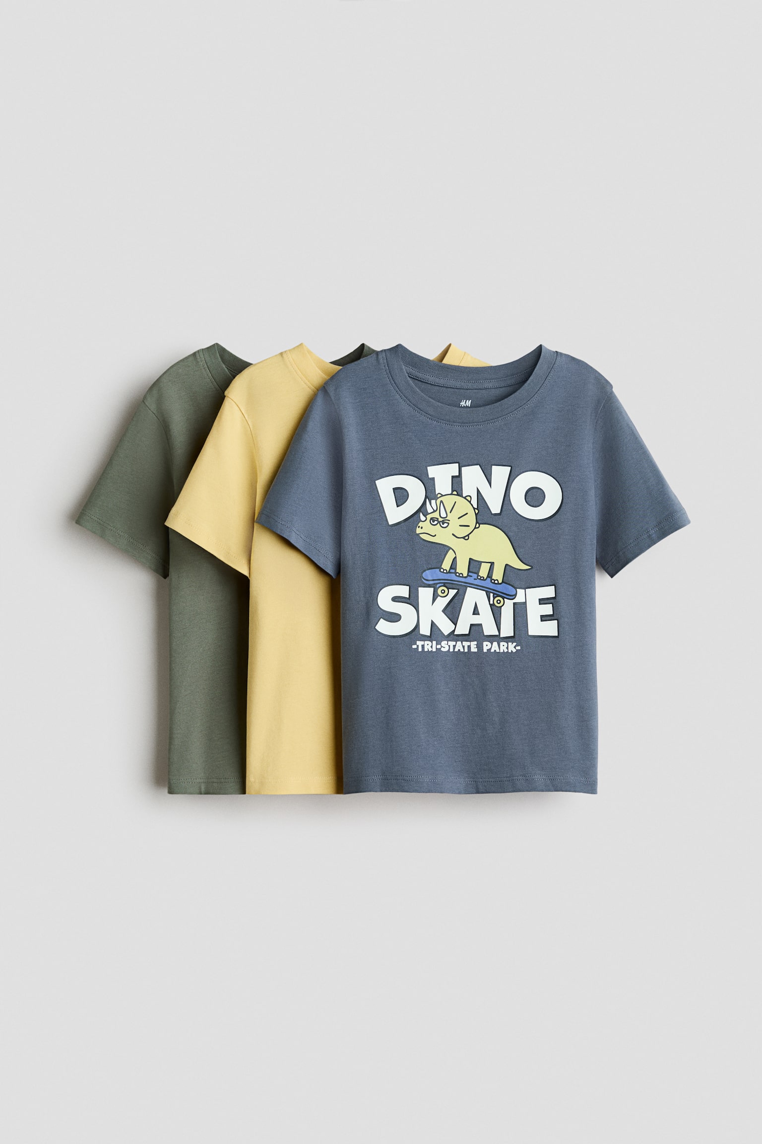 Three soft cotton jersey t-shirts are overlapping and facing front, showcasing a dark green tee in the back, a yellow tee in the middle, and a blue-grey tee in the front. The front tee is displaying a cartoon graphic of a dinosaur on a skateboard with the text "DINO SKATE," while all three are featuring a round, rib-trimmed neckline and short sleeves.