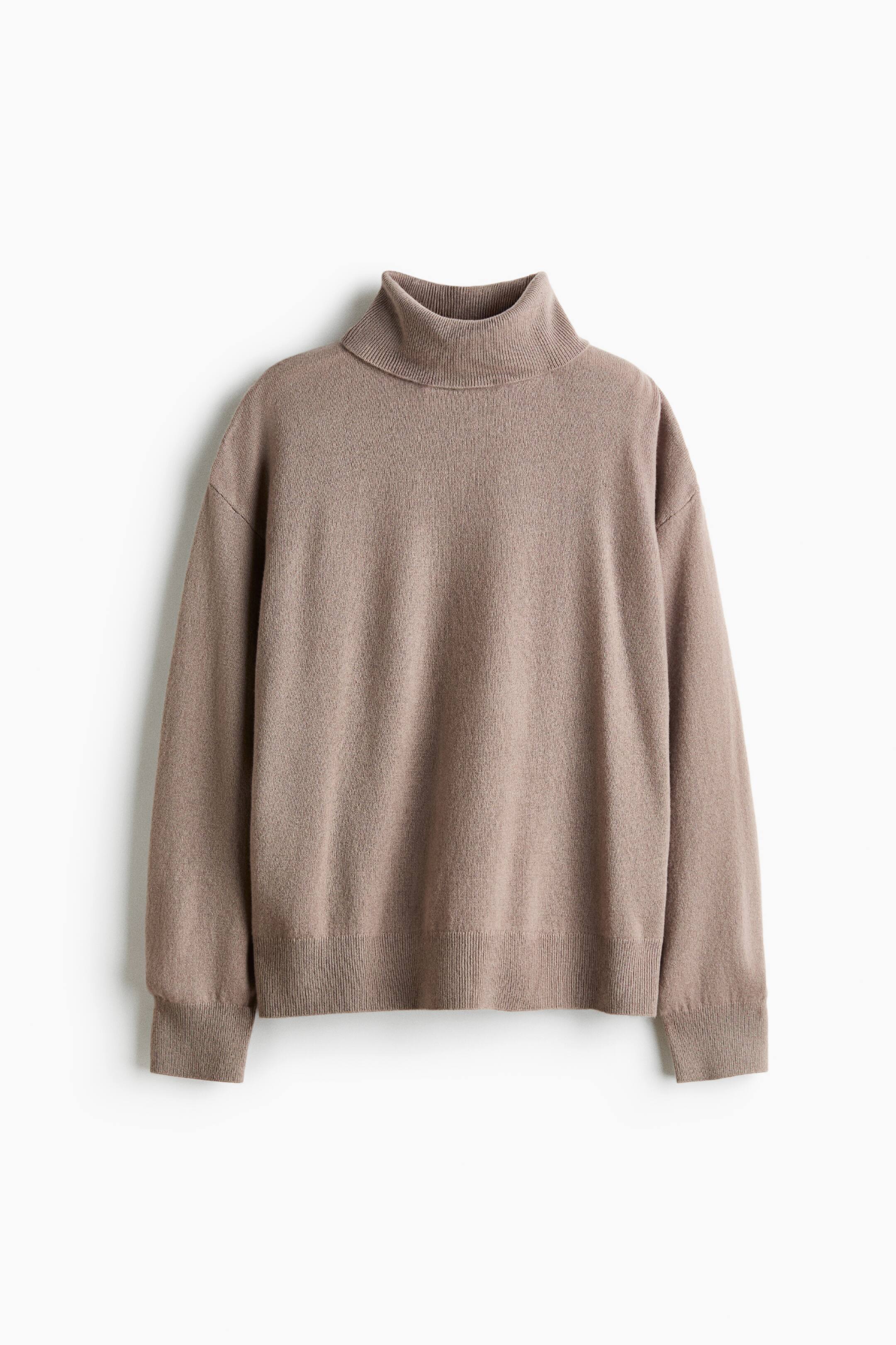 Ver imagen más grande: Un jersey de cachemir de punto fino en topo cuelga en posición plana y orientado de frente, mostrando una silueta oversize con cuello alto de canalé, hombros caídos y mangas largas rematadas con puños de canalé y bajo de canalé.