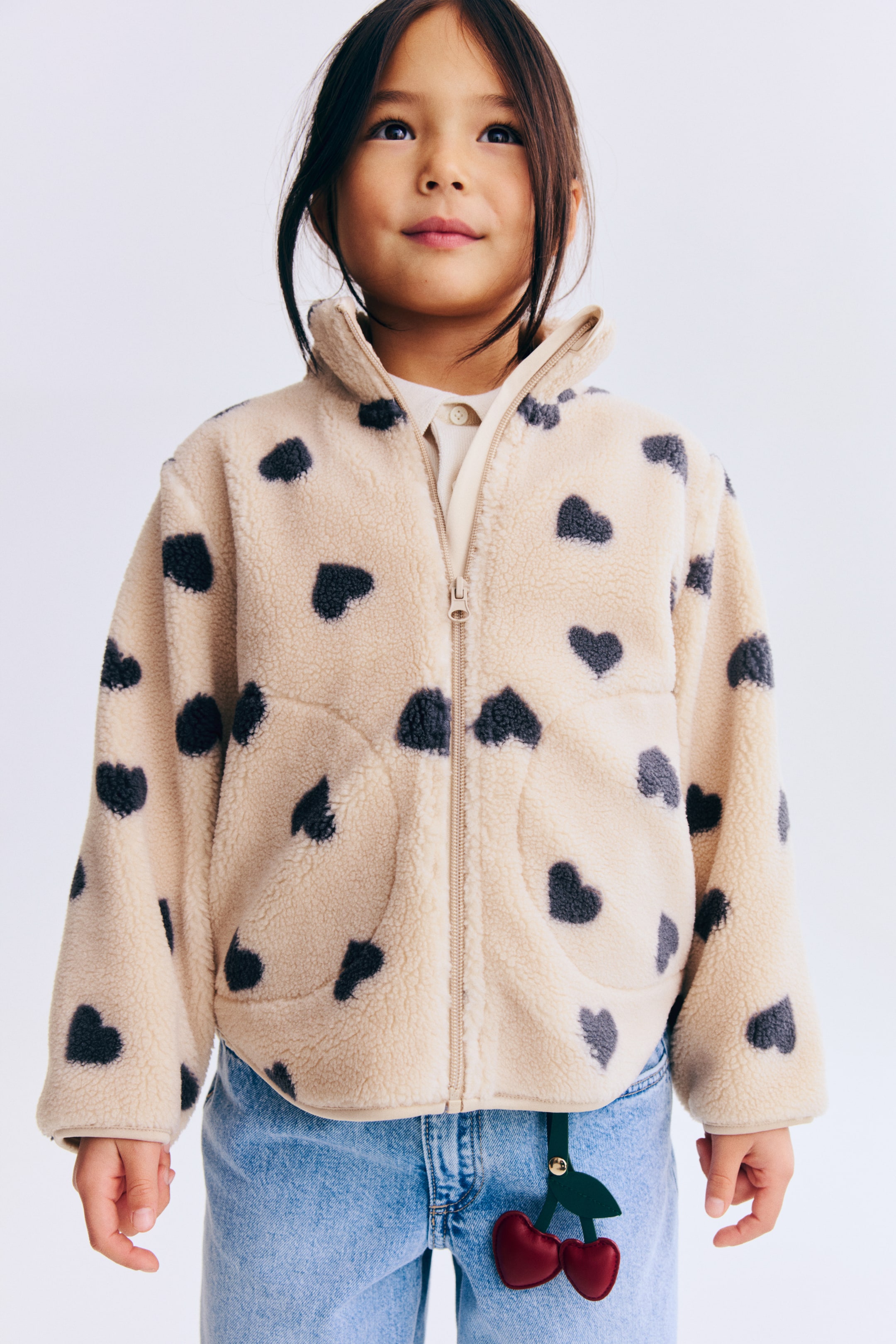 View larger image: A beige fleece jacket features scattered dark‑grey heart patterns across its surface. The front has a full‑zip closure, a stand‑up collar and two discreet vertical‑entry pockets. It is shown worn over a light‑coloured collared polo shirt.