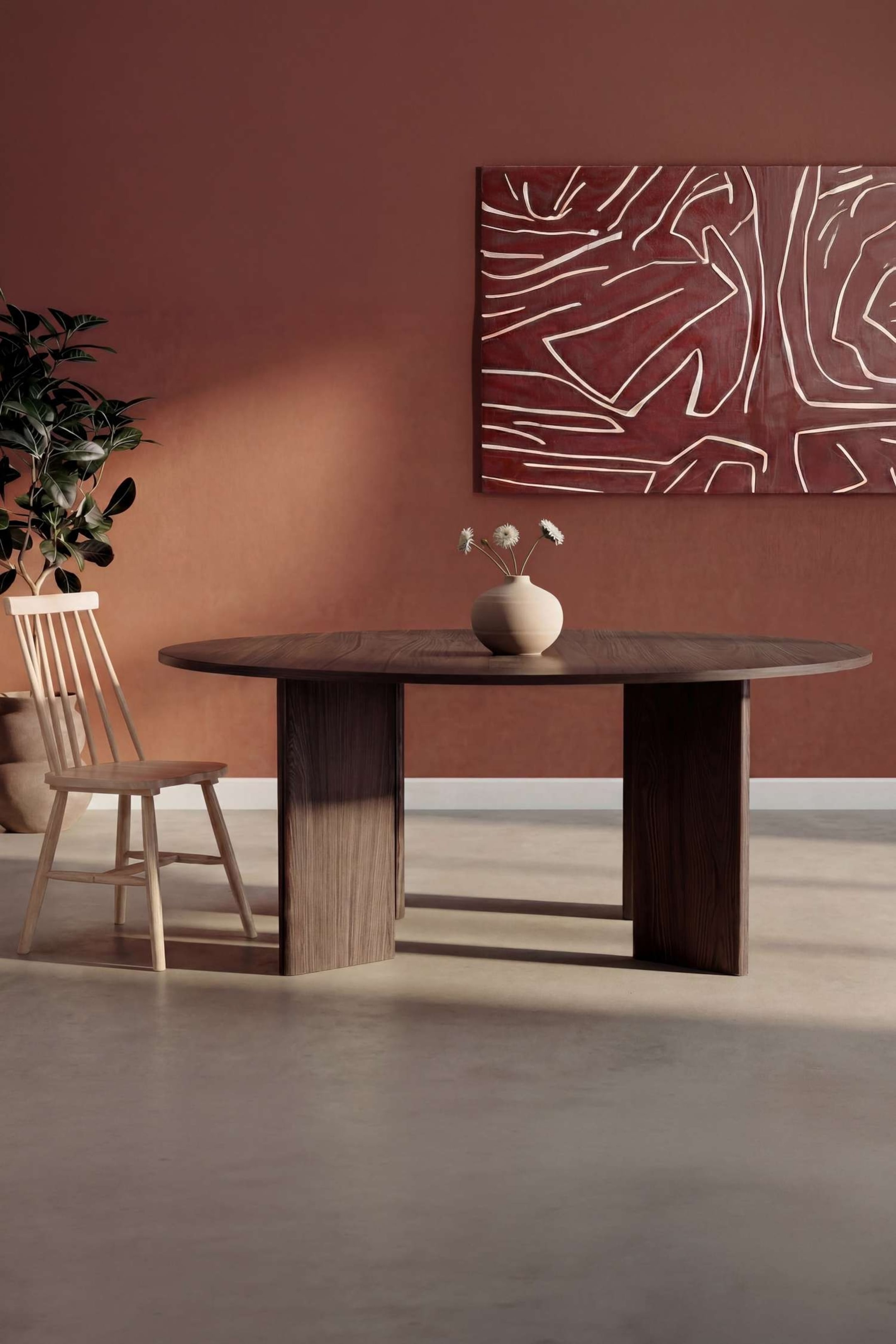Agrandir l'image: La table à manger en bois brun foncé est de forme ovale, présentant un veinage du bois bien visible sur sa surface supérieure, et elle est soutenue par deux piètements rectangulaires épais. Un vase en céramique de couleur claire contenant trois fleurs blanches est placé au centre de la table.
