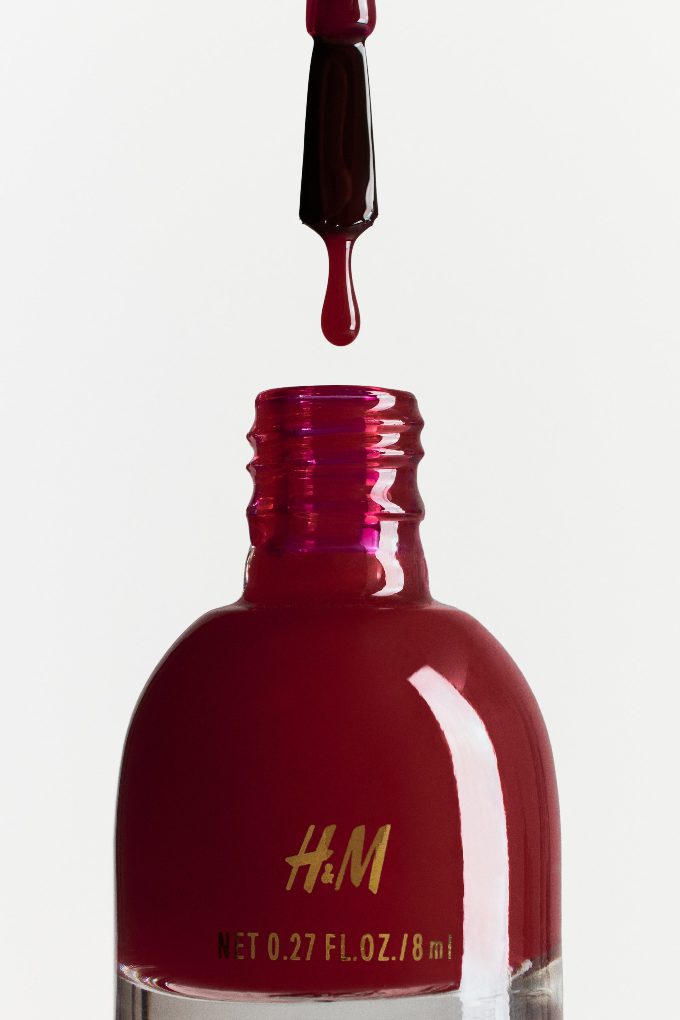 Grösseres Bild ansehen: Eine klare Glasflasche mit Nagellack ist mit tiefrotem Lack gefüllt und zeigt das "H&M"-Logo sowie das Produktvolumen auf der Vorderseite. Ein Applikatorpinsel, der mit demselben roten Lack beschichtet ist, befindet sich über der Flasche, während ein Tropfen Lack in Richtung der Gewindeöffnung fällt.