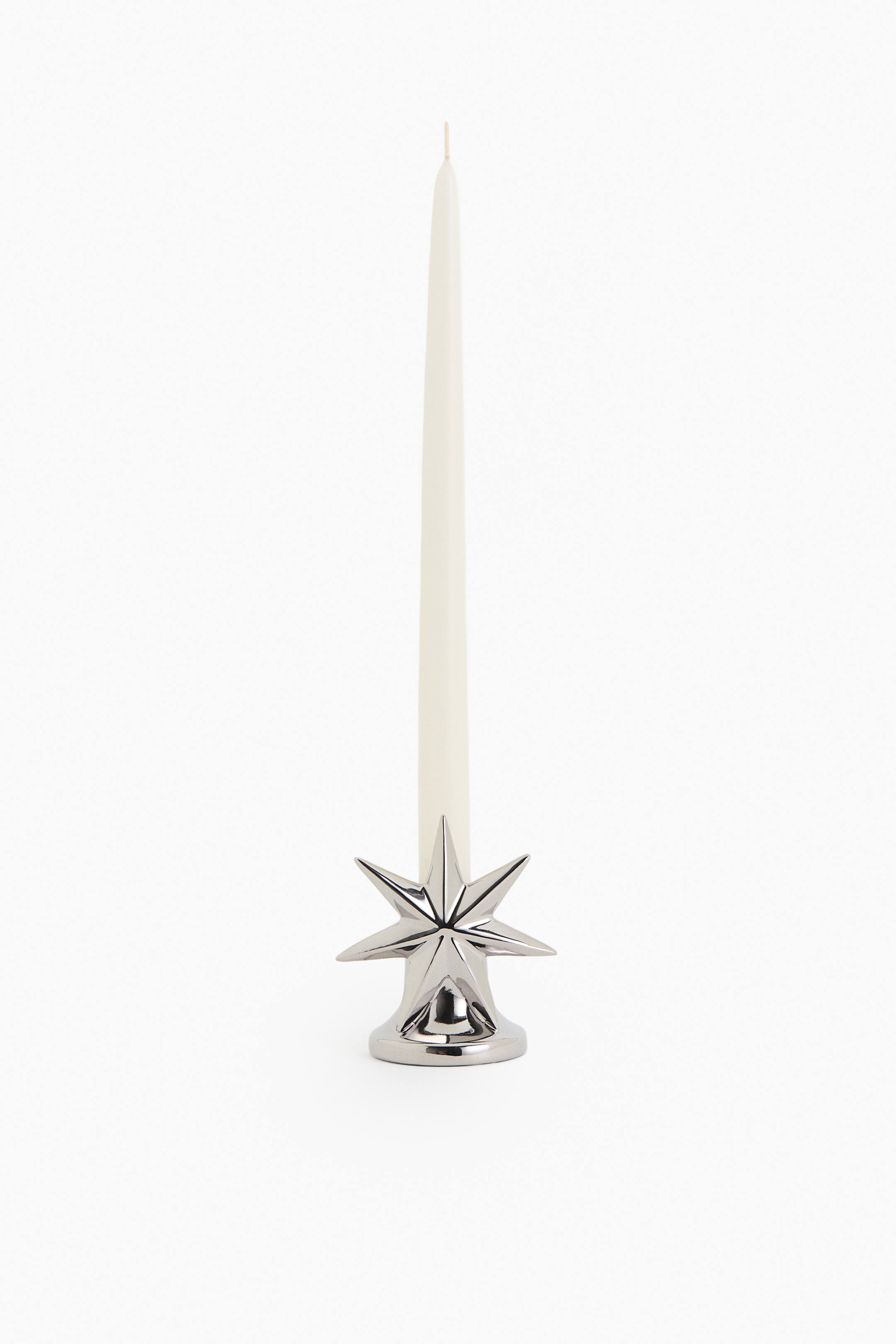 View larger image: A cream-colored tapered candle stands upright in a shiny silver starburst-shaped candle holder, facing forward.